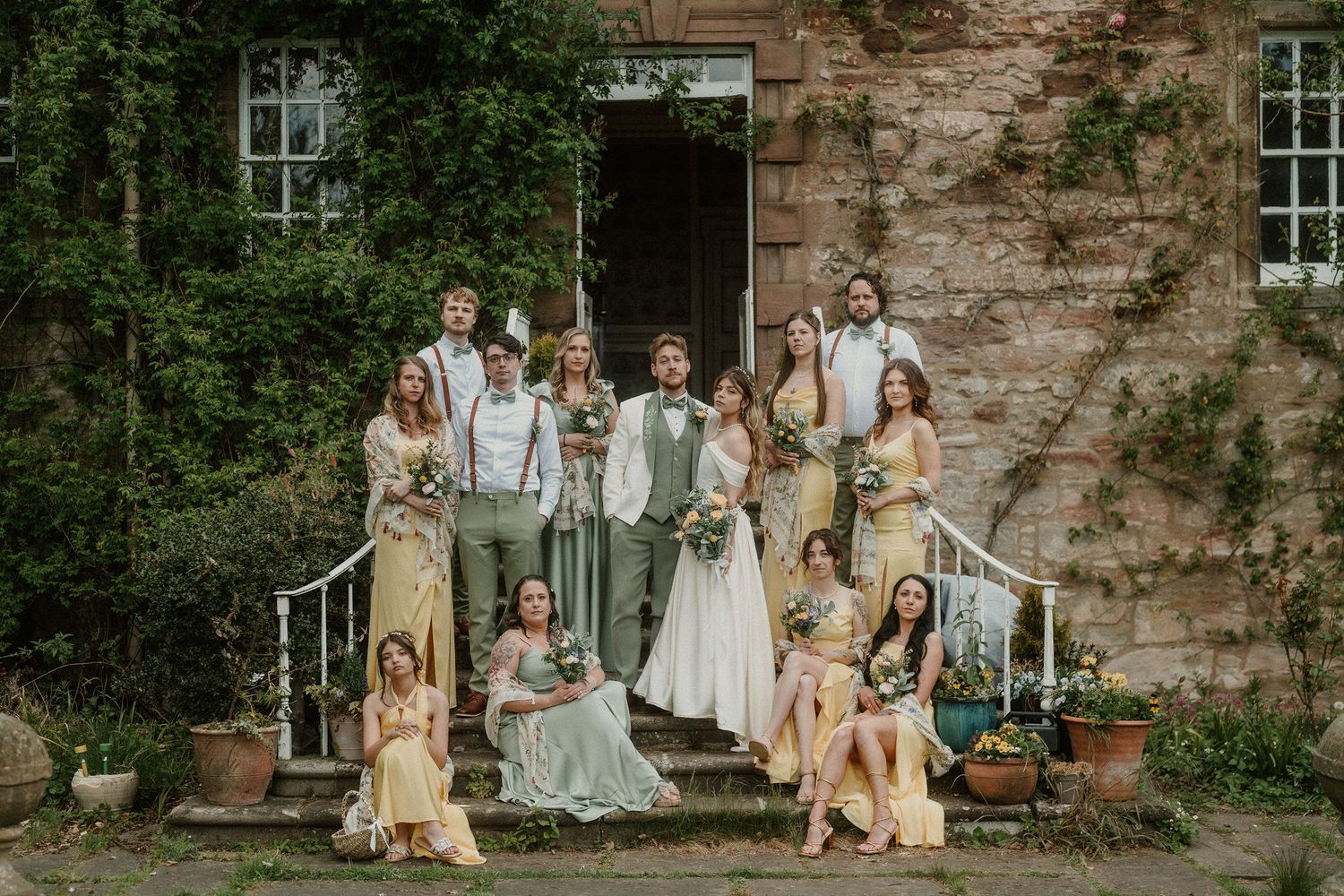 Wedding party poses on stone steps of rustic ivy-covered venue, bridesmaids in sage green and yellow dresses with bouquets.