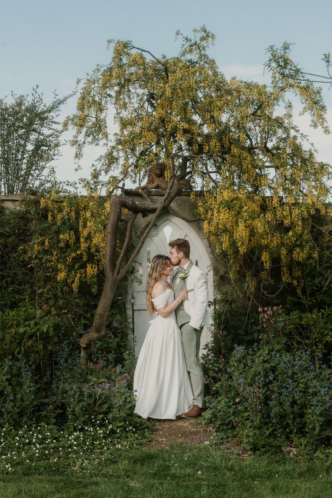 Bride and groom embracing under a flowering weeping tree arch in a lush garden wedding setting.