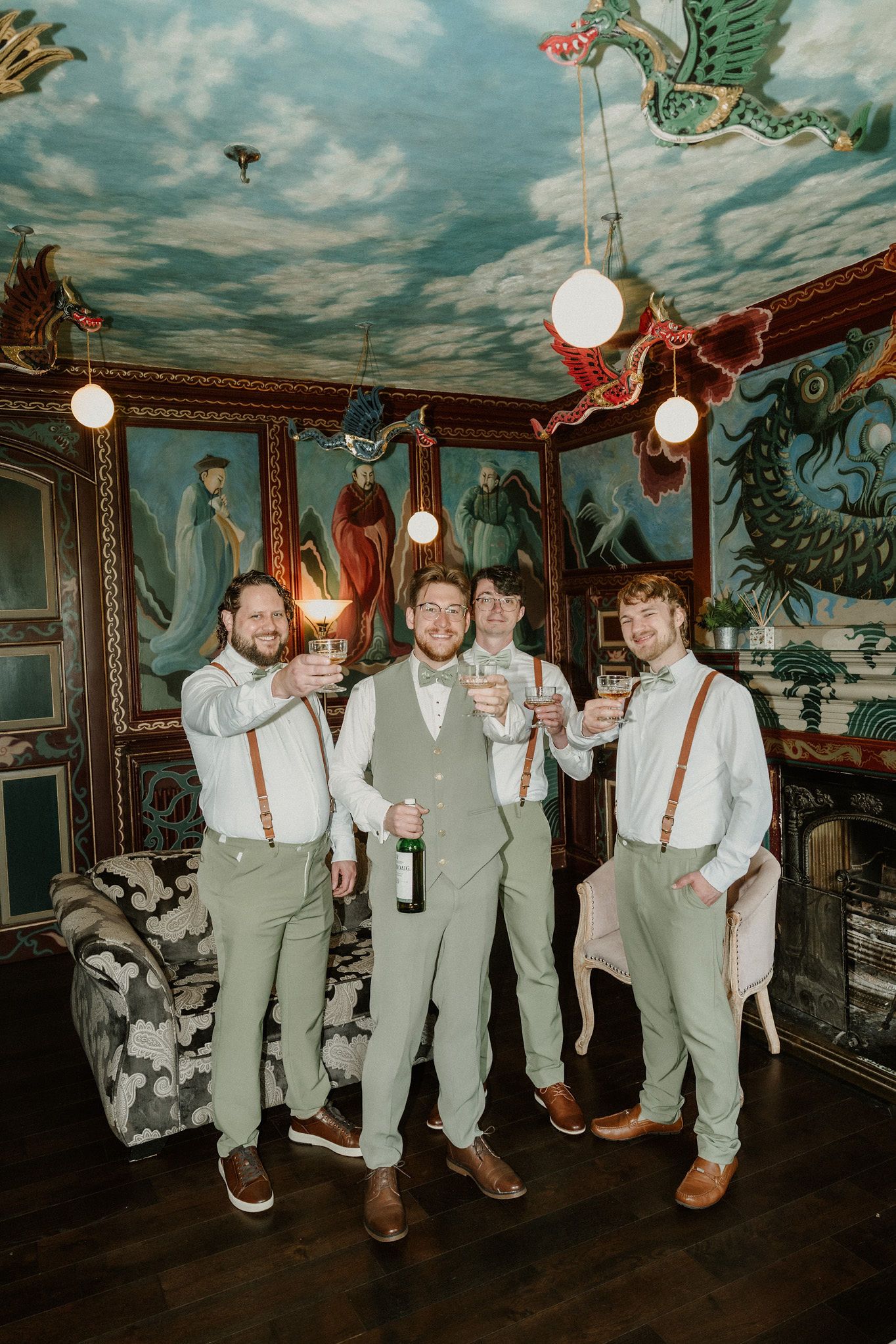 Groomsmen in sage green pants and suspenders toast with drinks in a vibrant, mural-covered room.