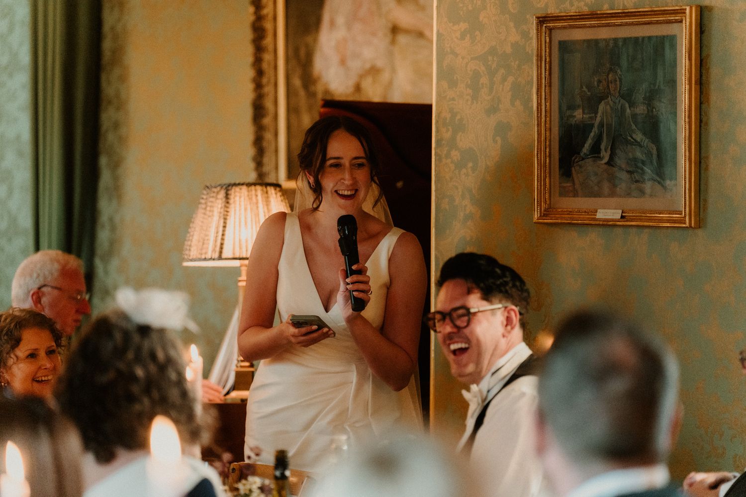 A smiling woman in a white dress holds a microphone and gives a speech while seated wedding guests laugh in an elegant, candlelit room.