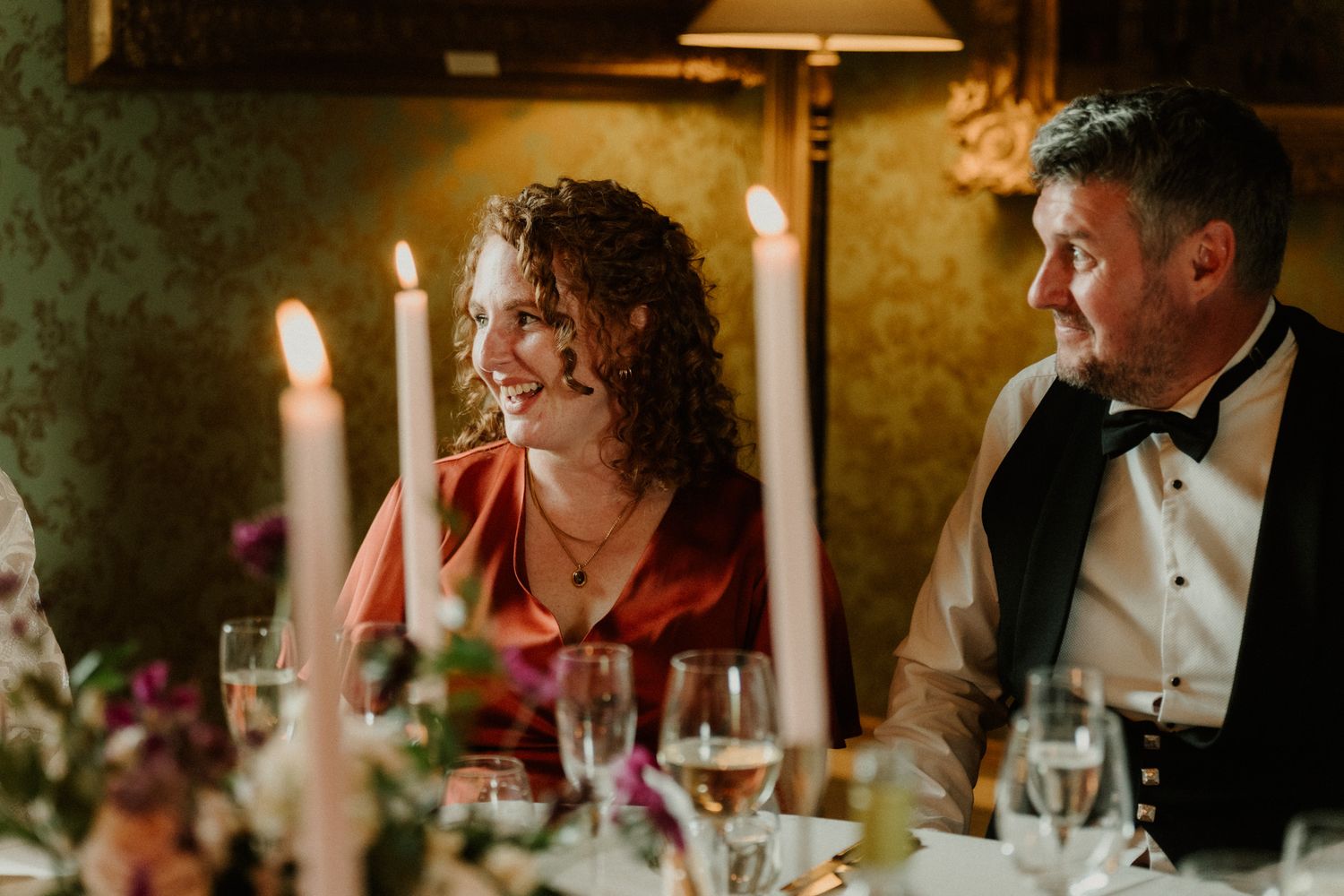 Smiling woman with curly hair and a man in a bow tie and vest sit at a candlelit table with wine glasses and flowers.