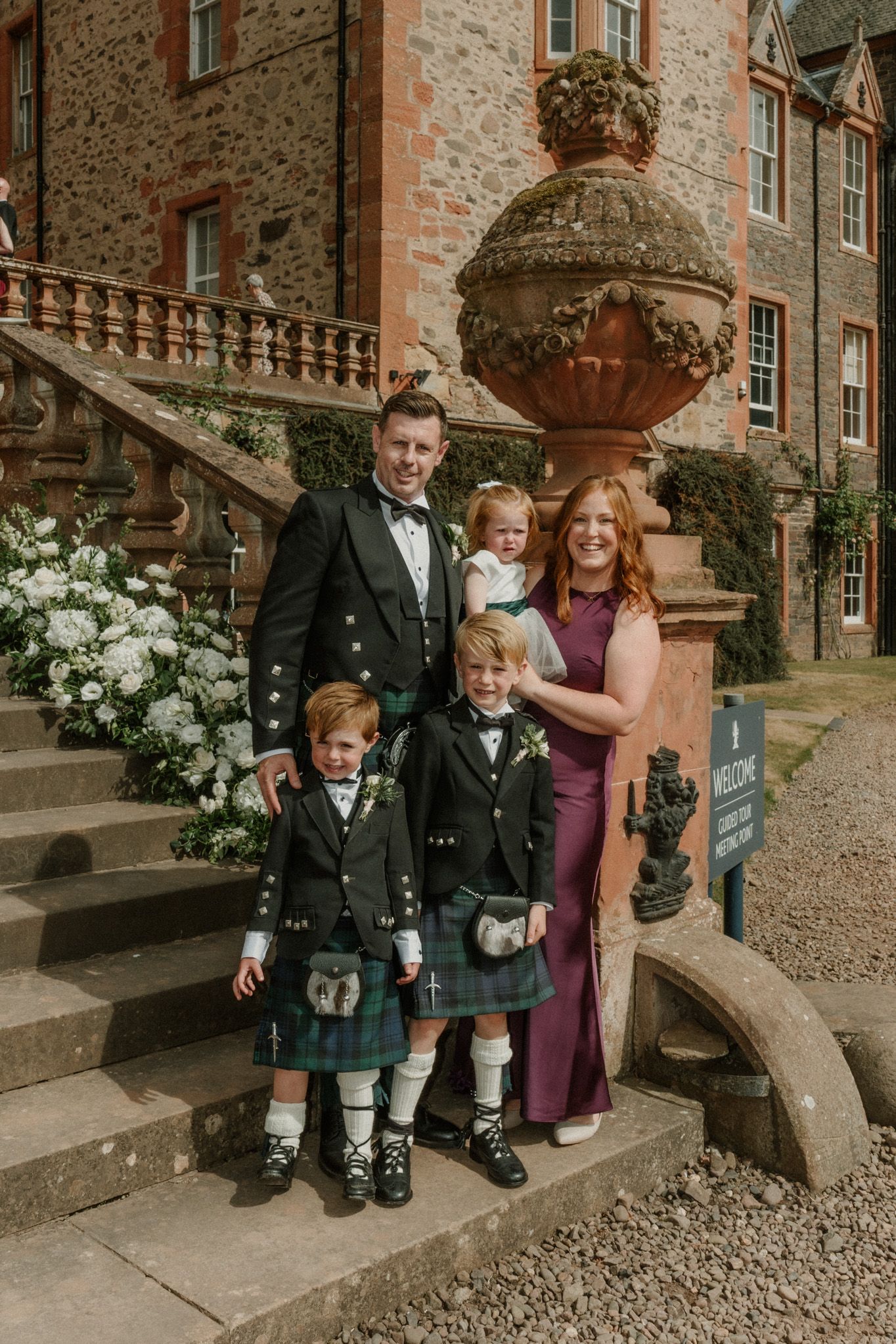 Family in Scottish wedding attire with two boys in kilts and a toddler pose on stone steps beside white flowers at a historic castle.