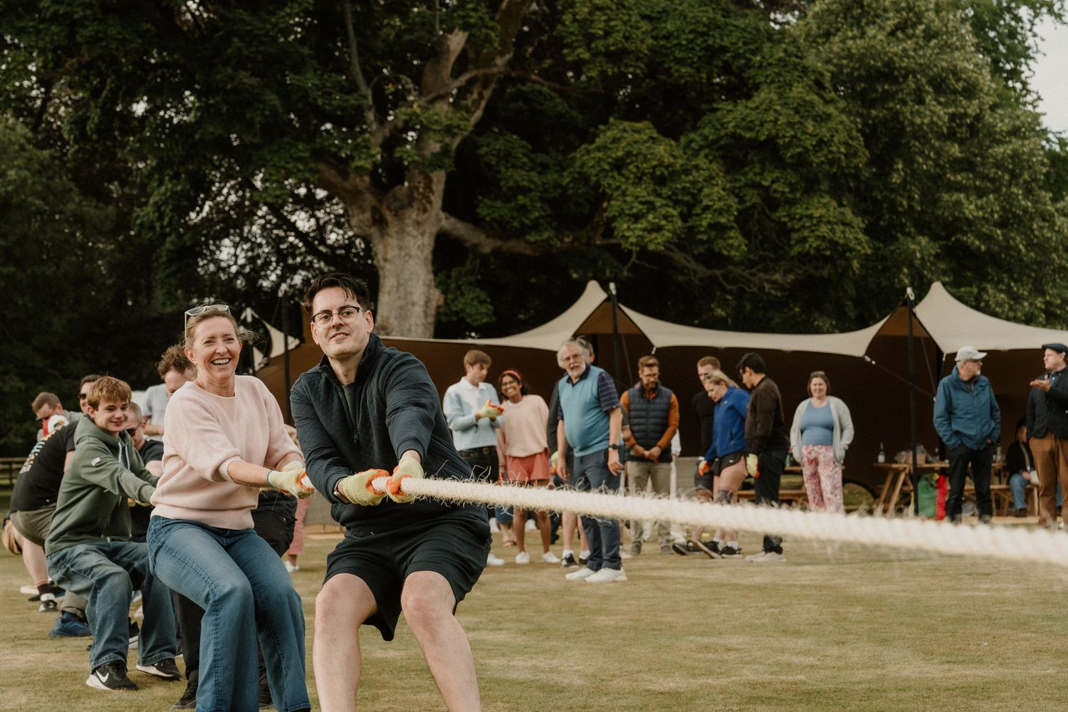 Group of people playing tug of war on a grassy lawn with spectators and open-sided tents under trees in the background.