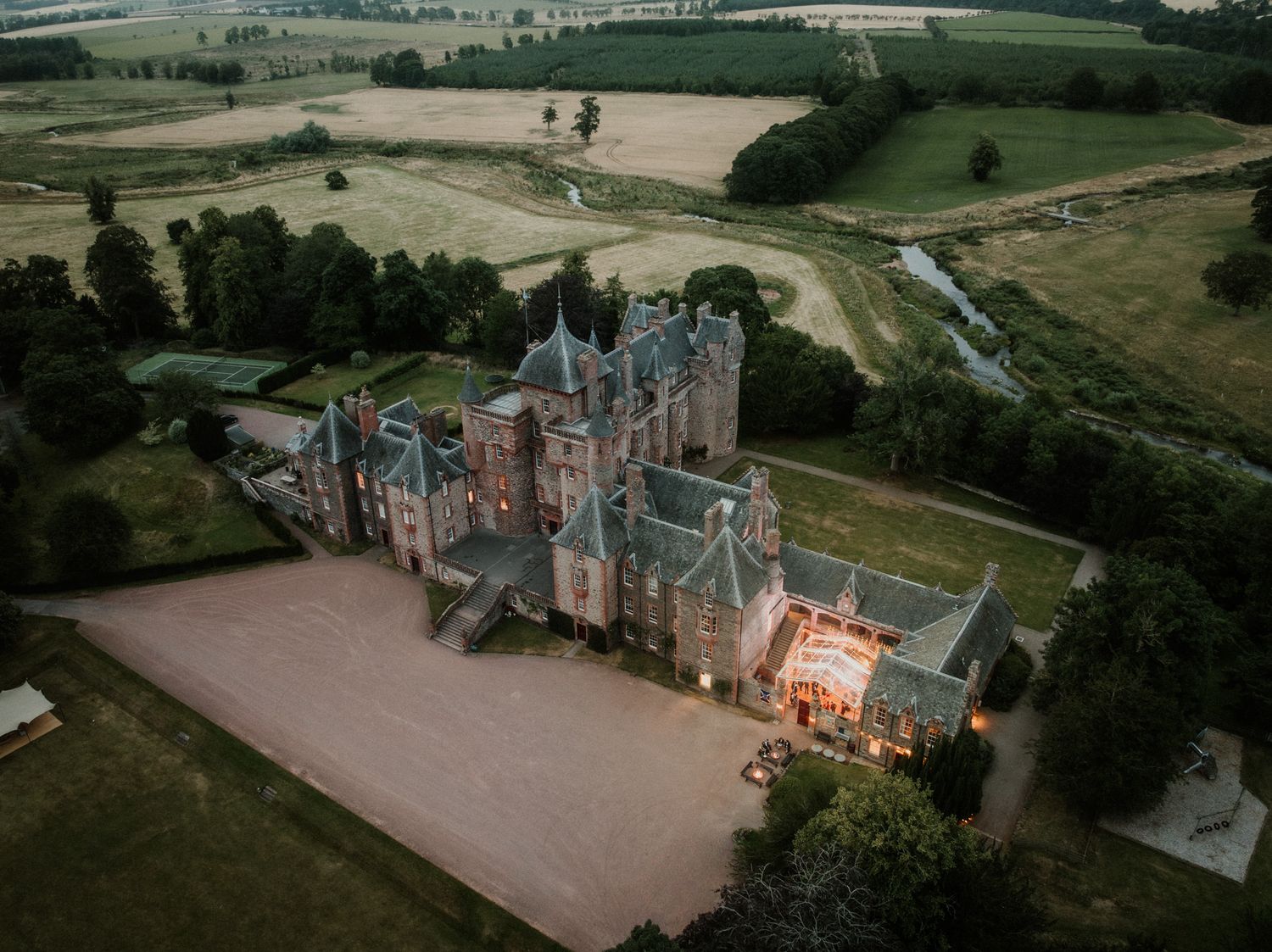 Aerial view of a historic stone castle with turrets, a lit entrance, gravel courtyard, and surrounding woodland and countryside at dusk.