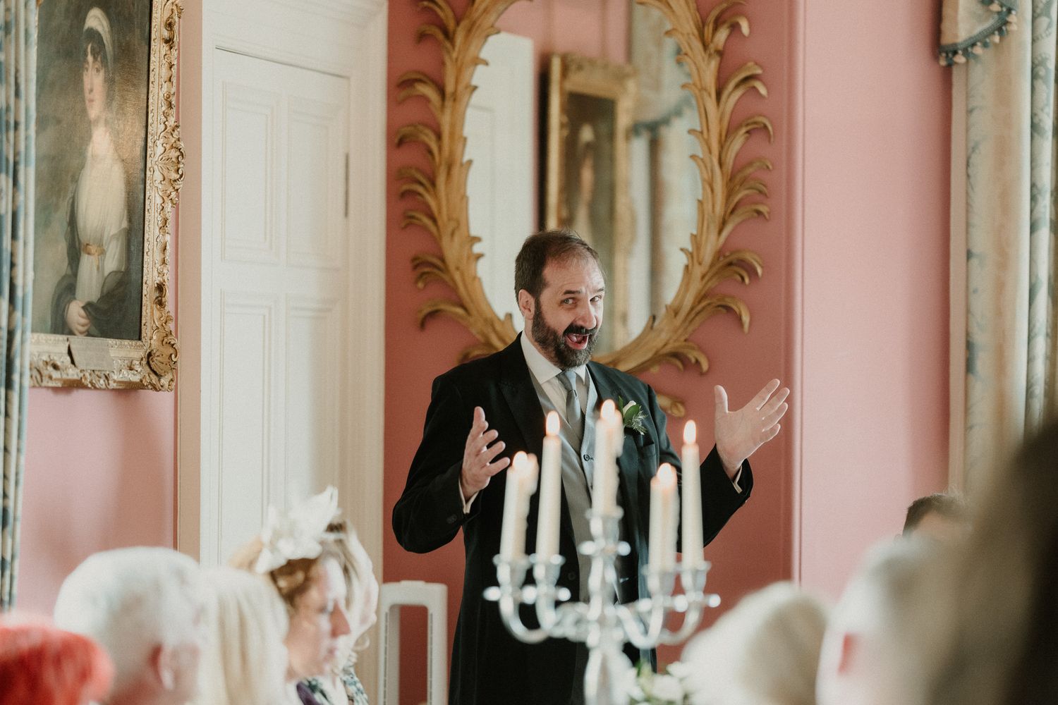 A bearded man in a suit giving a speech at an elegant wedding reception in a pink ornate room.