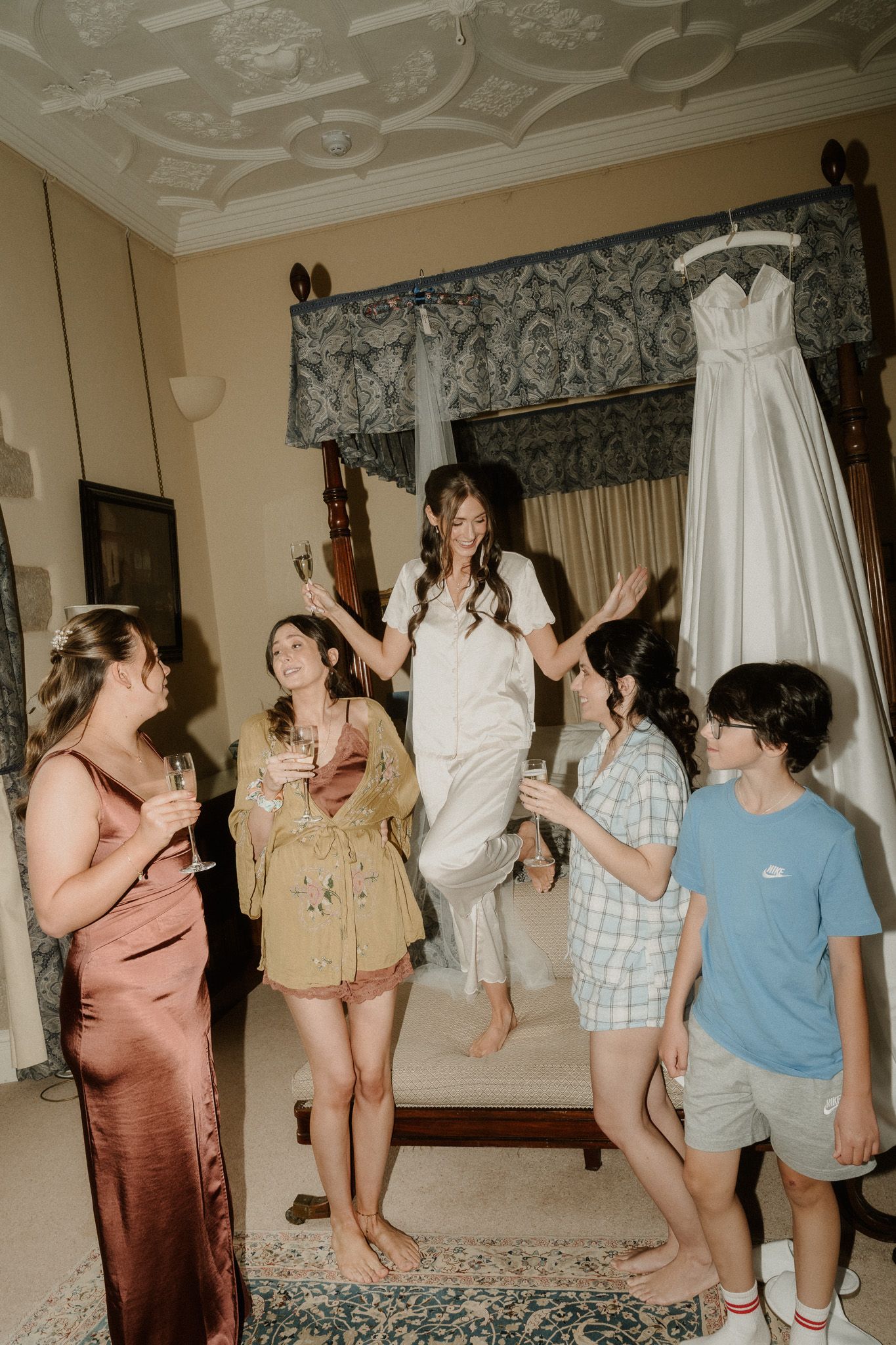 Bride jumping for joy with bridesmaids and guests in a elegant room with wedding dress hanging in background.