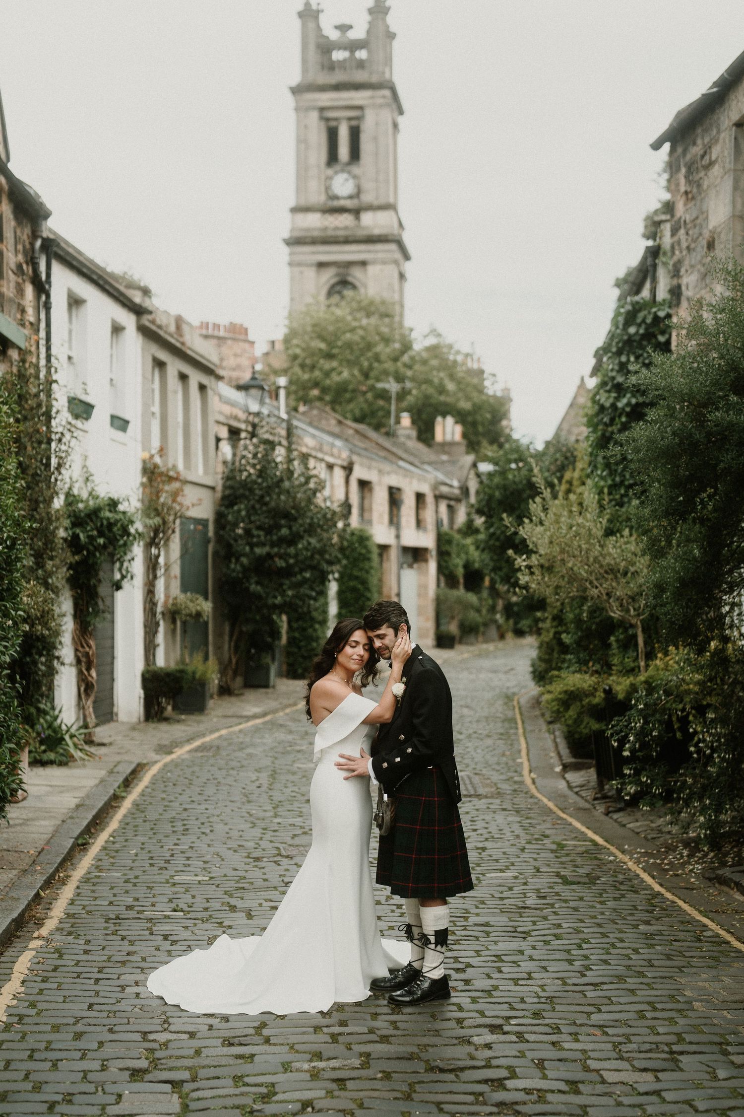 Romantic wedding couple embraces on scenic cobblestone street with historic church tower in background.