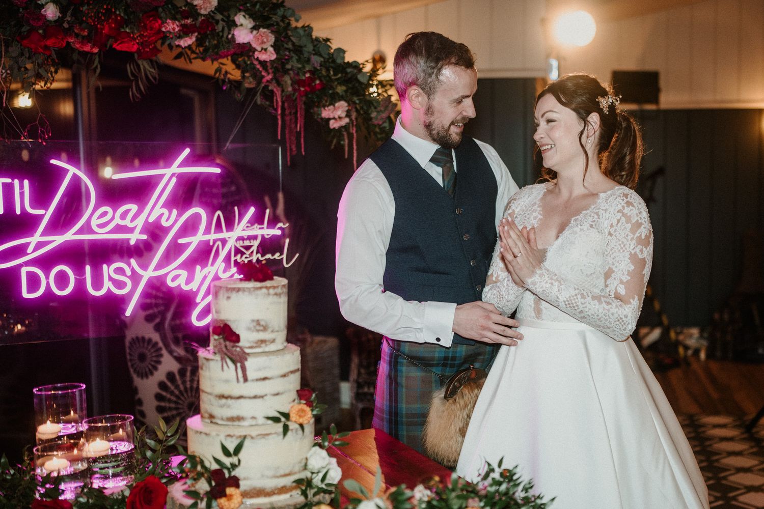 Wedding cake display with pink neon lighting and floral decorations at reception venue.