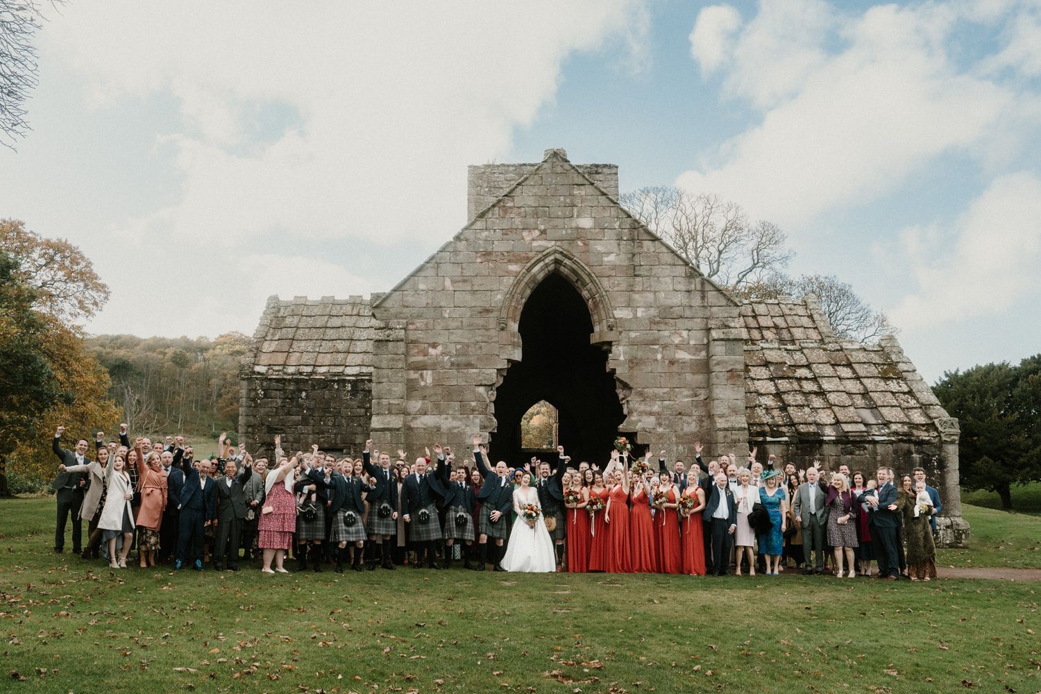 Large group wedding photo taken in front of historic stone building with arched entrance.