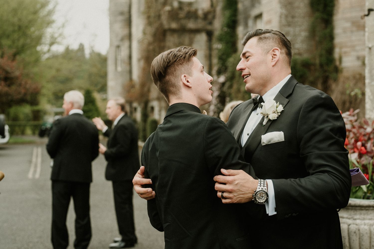 Wedding party members share embraces outside historic stone building.