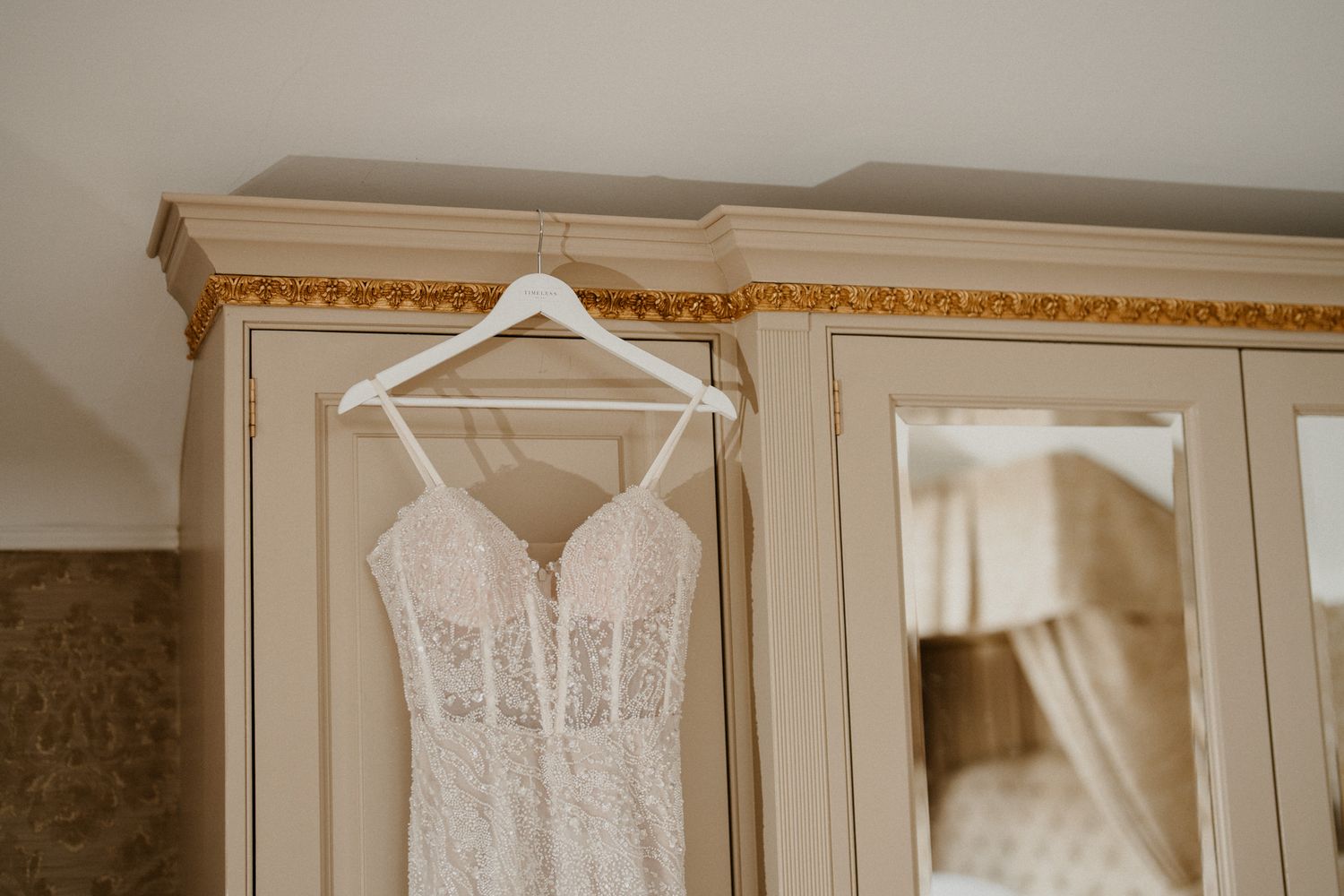 White lace wedding dress displayed on a hanger against cream-colored walls.