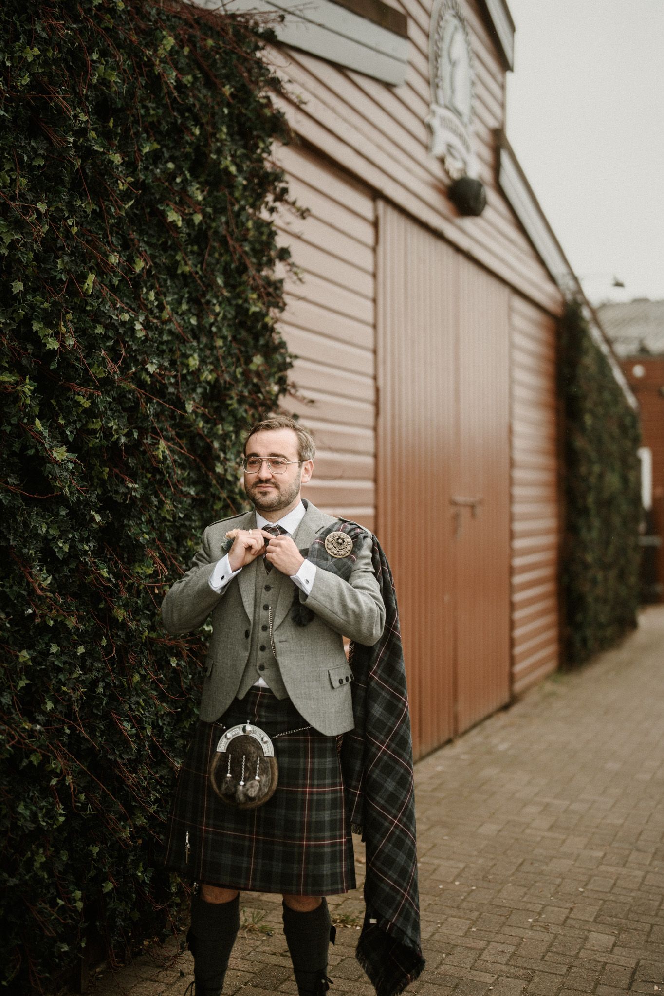 Man in traditional Scottish kilt and formal wear poses against rustic background.