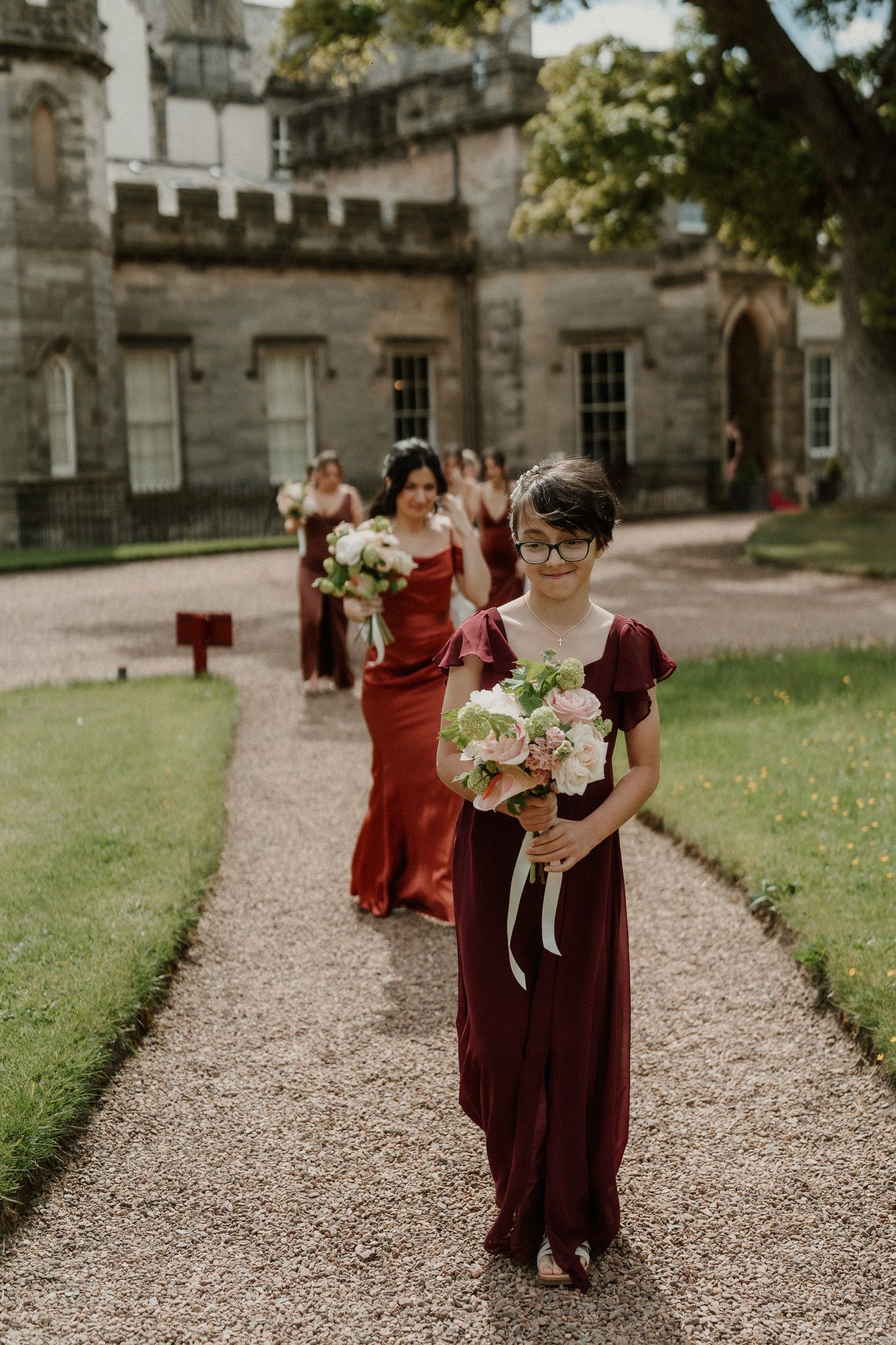 Bridesmaids in burgundy dresses walk along a gravel path carrying pink bouquets outside a historic stone venue.