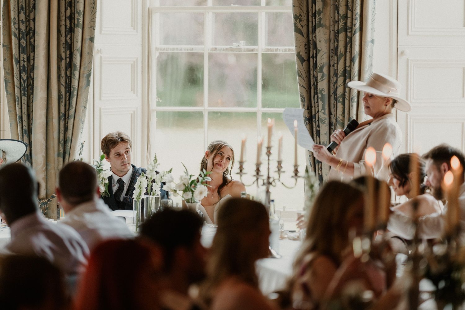 Bride and groom seated at wedding reception head table as guest in white hat delivers a heartfelt speech.
