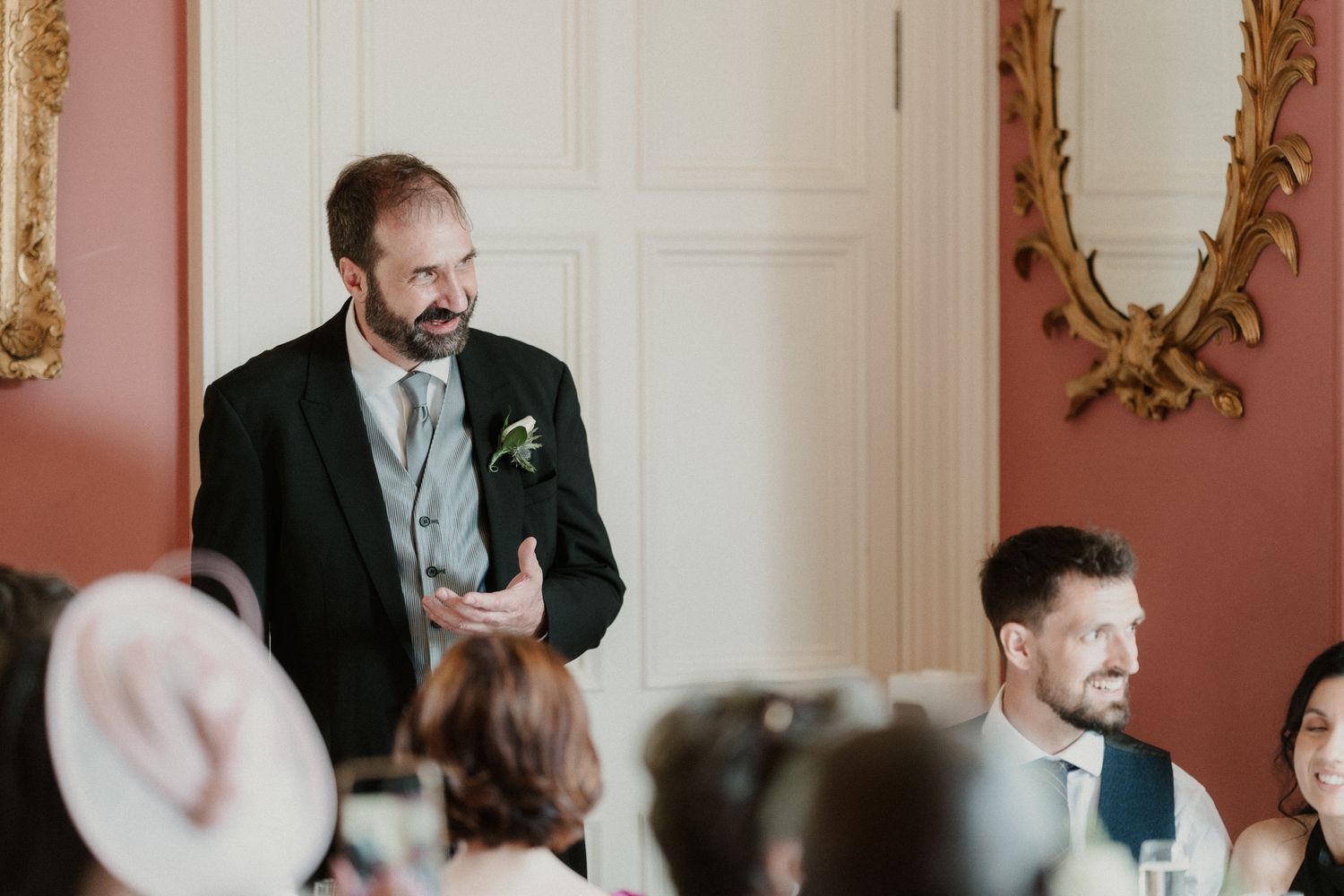 Man in black suit giving a speech at an elegant wedding reception in a ornate room with gold mirrors.