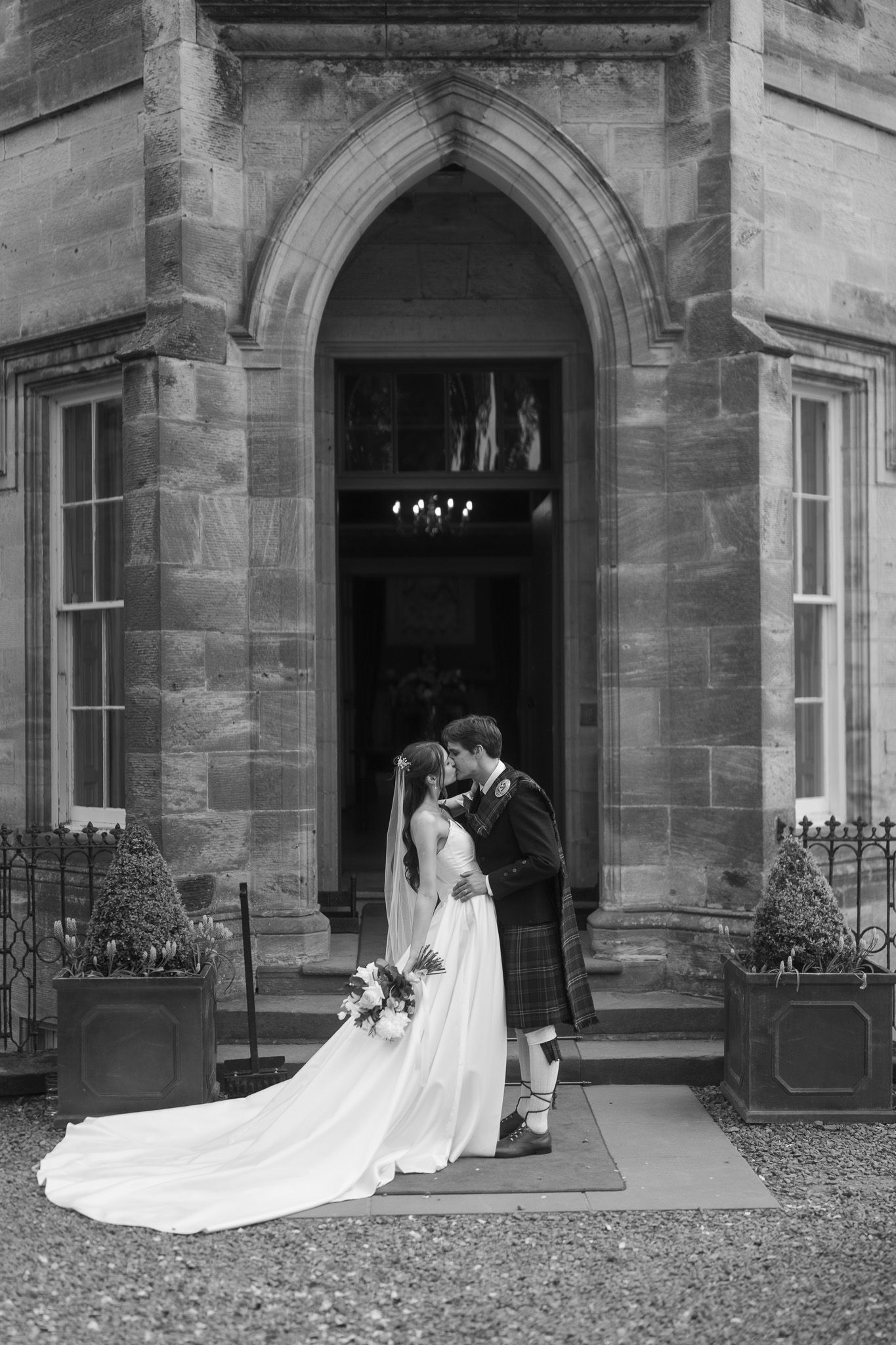 Black and white photo of a bride and groom kissing in a Gothic stone archway, bride in flowing gown with long veil.
