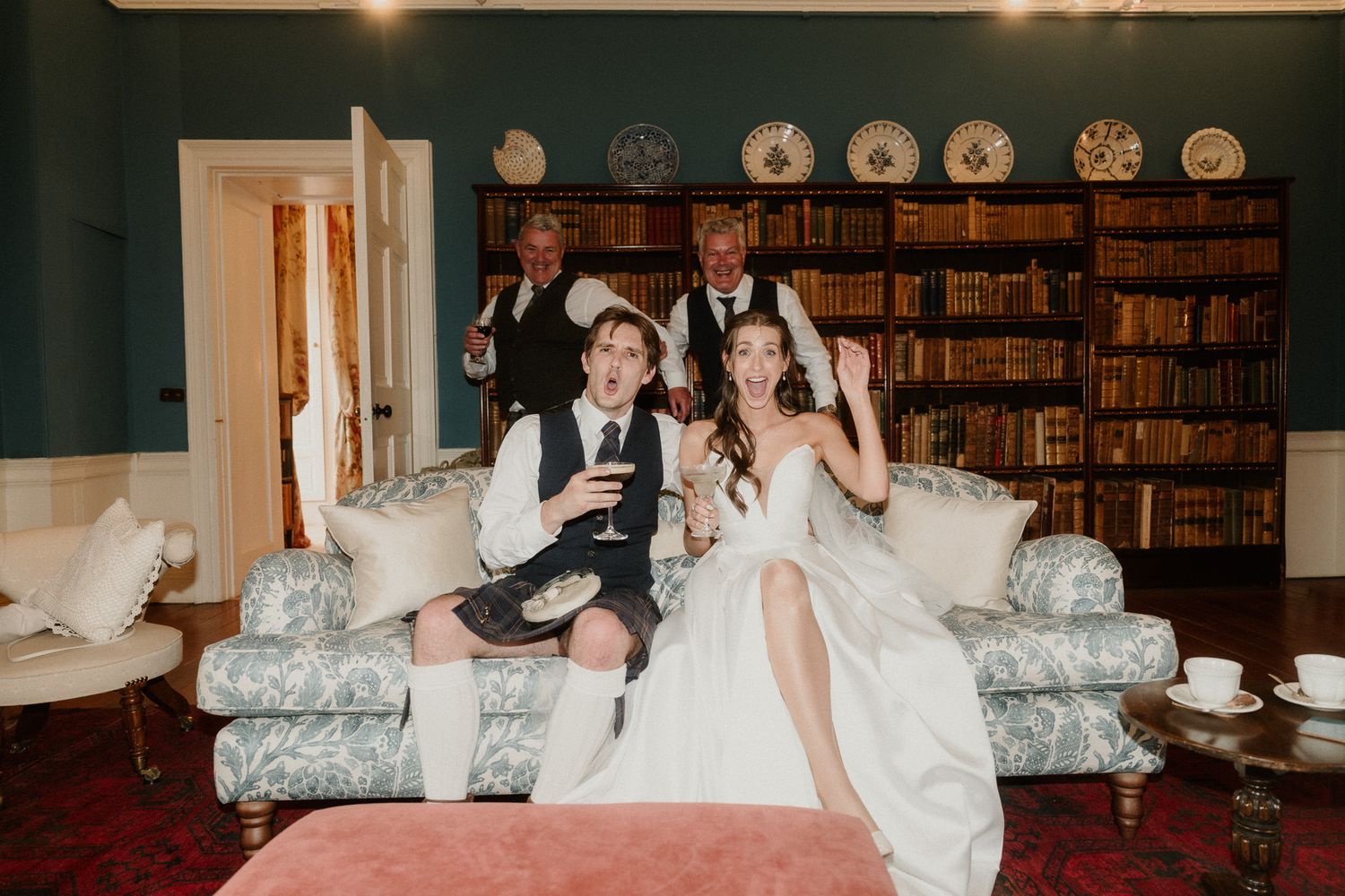 Bride and groom celebrate with guests on a floral sofa in an elegant library room during their wedding reception.