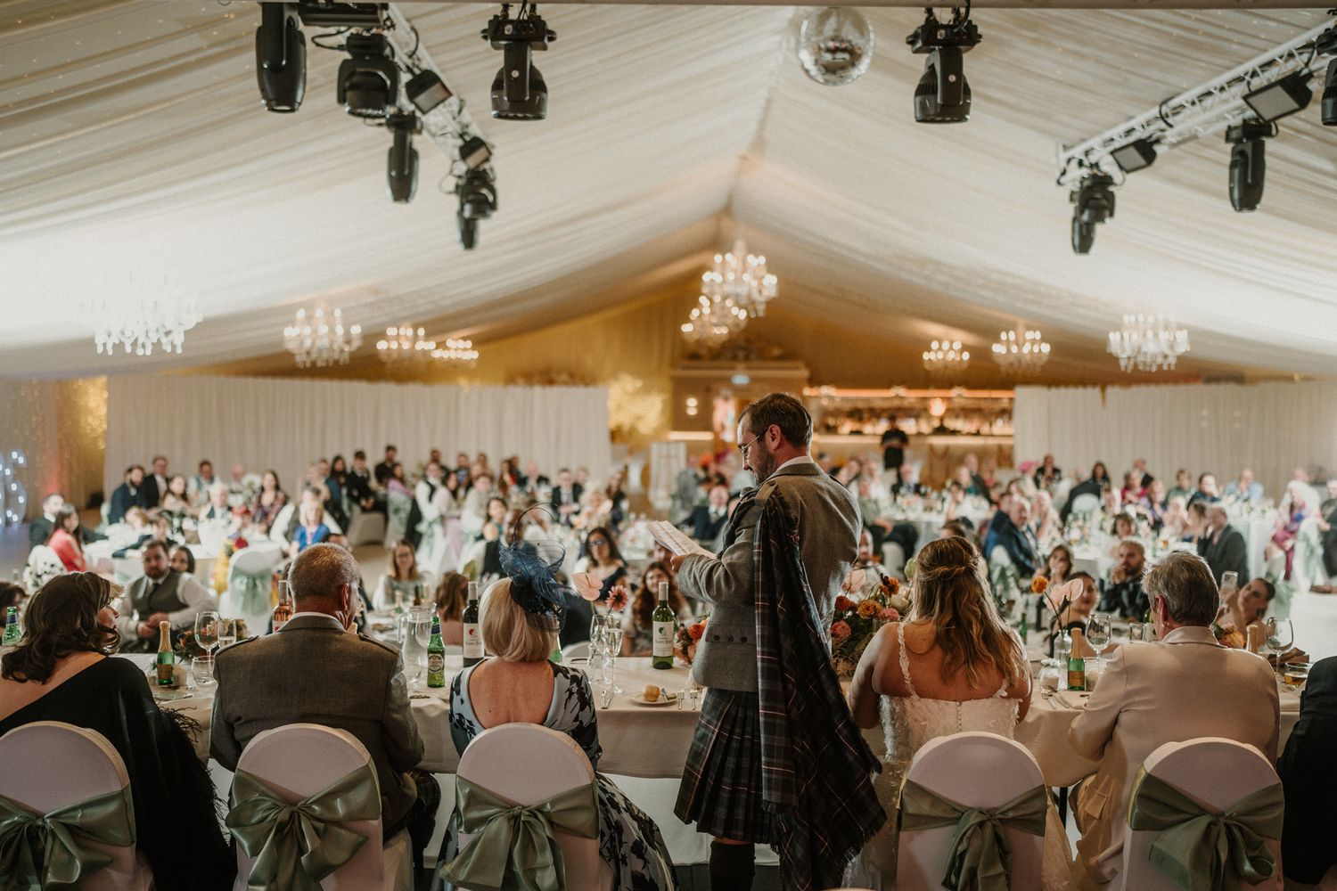 A wedding reception venue with draped ceiling, string lights and guests seated at round tables.