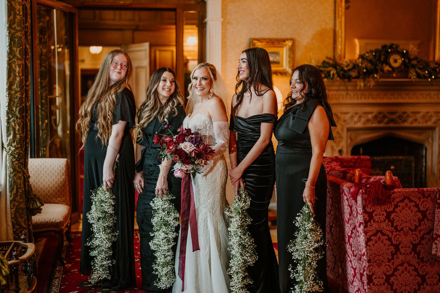 Wedding party in elegant formal attire gathered in an ornate room with red decor and floral arrangements.