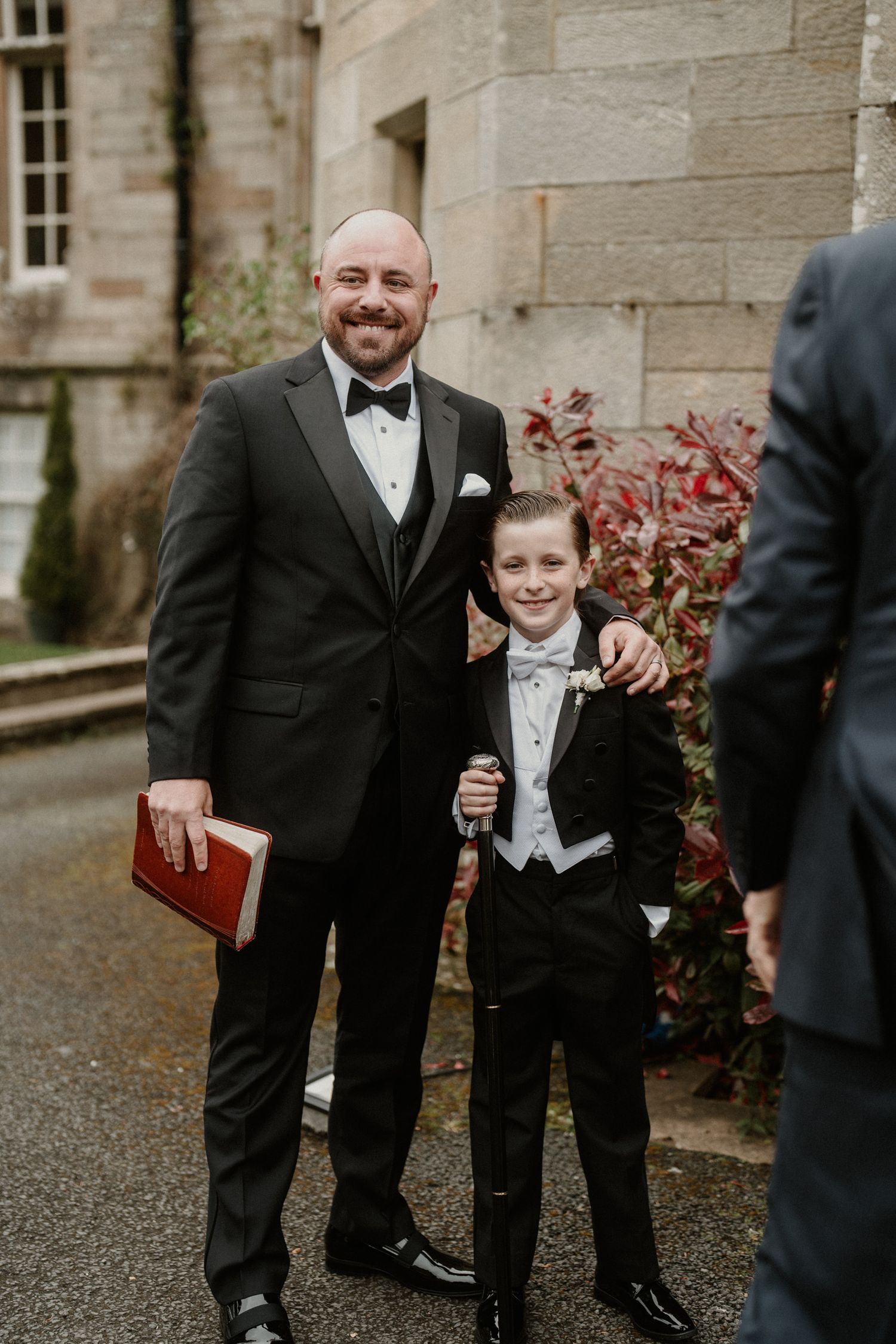 Wedding party members in formal black tuxedos gather outside a stone building.