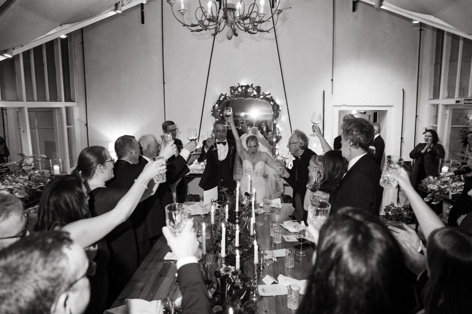 Wedding guests raise their glasses in celebration during an indoor reception with elegant chandeliers.