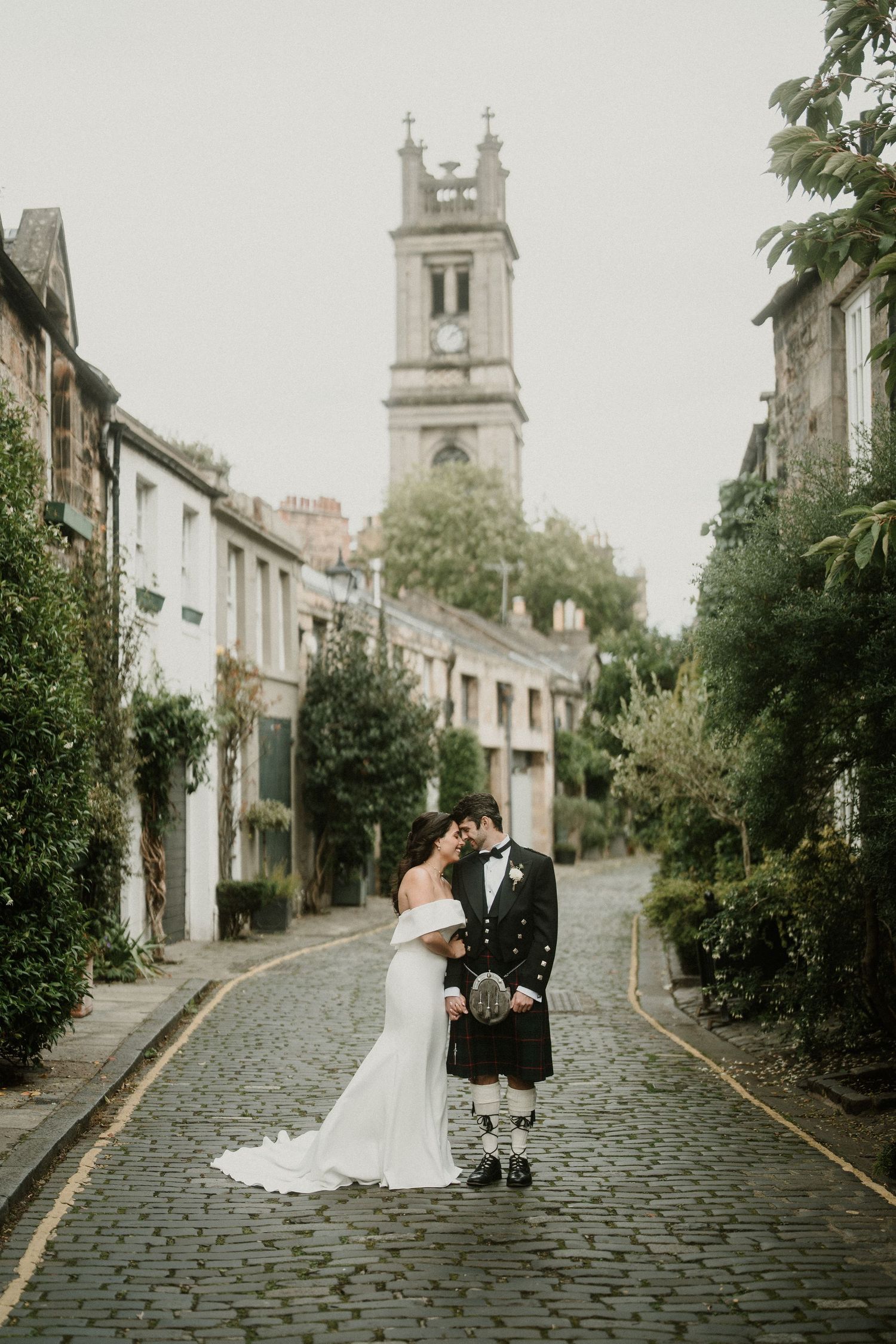Couple in wedding attire and traditional Scottish dress pose on historic street.