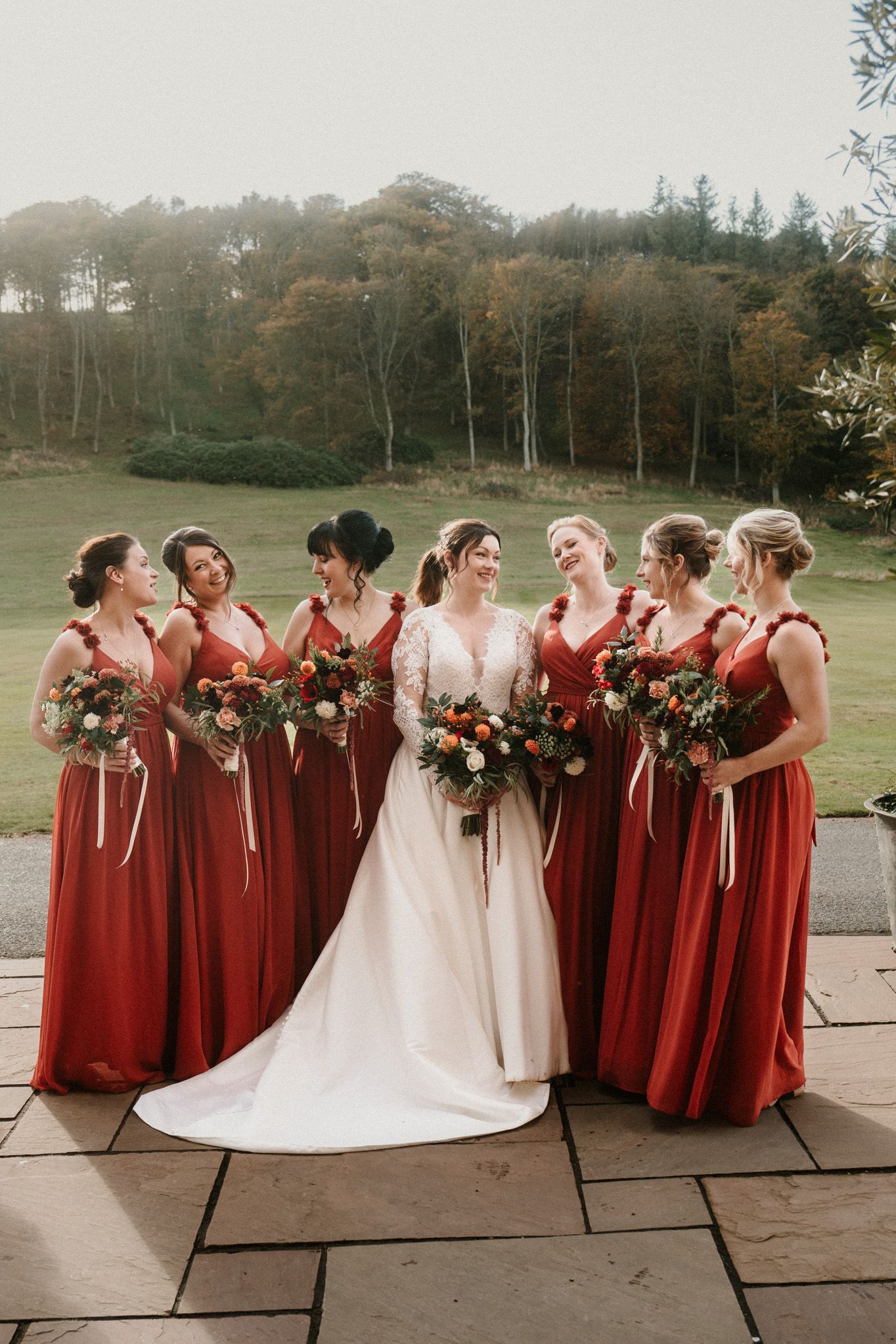 Bridesmaids in burgundy dresses gather around bride in white gown on stone patio.