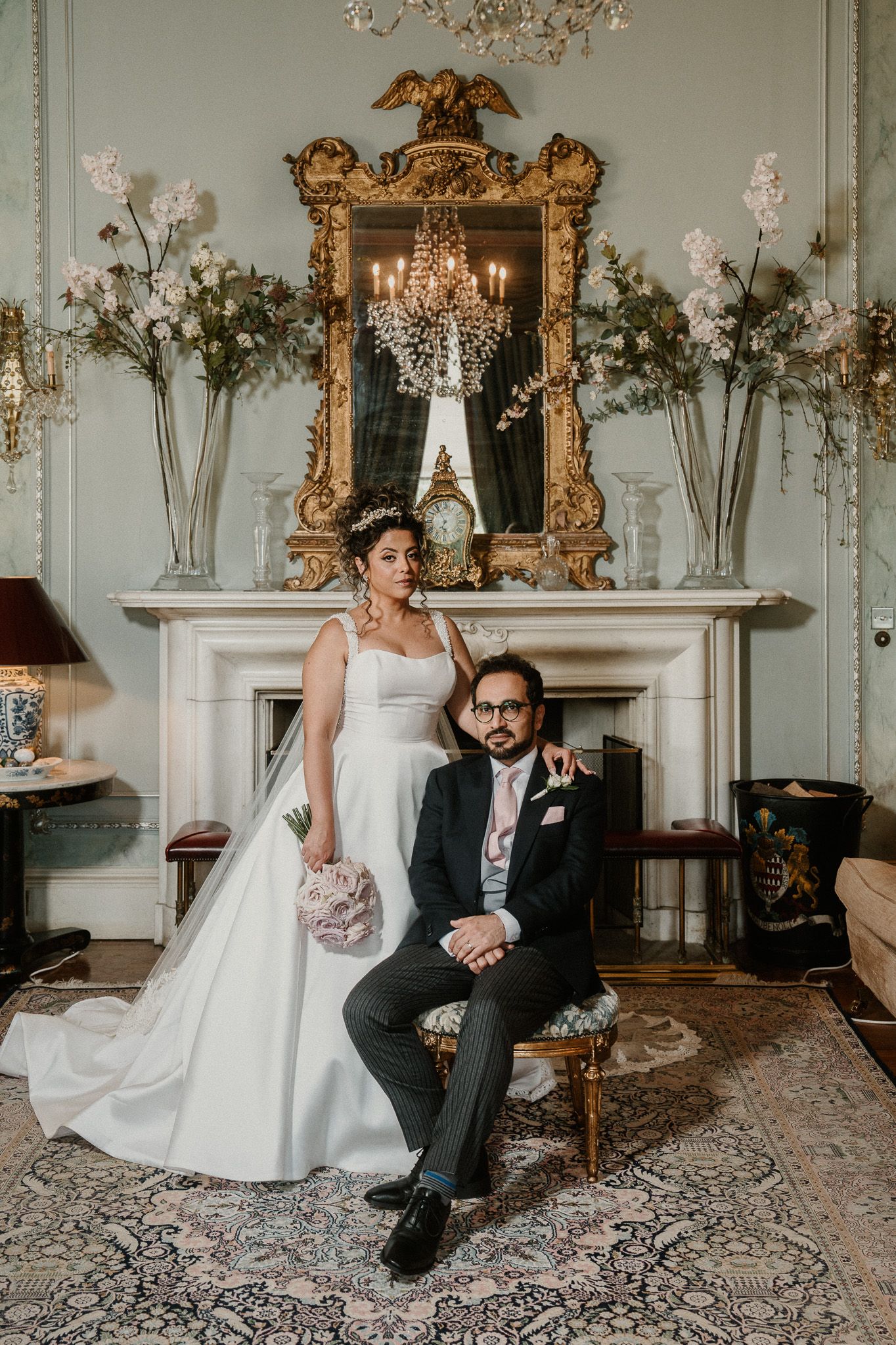 Wedding couple poses elegantly in ornate room with gilded mirrors and chandelier during formal portrait session.