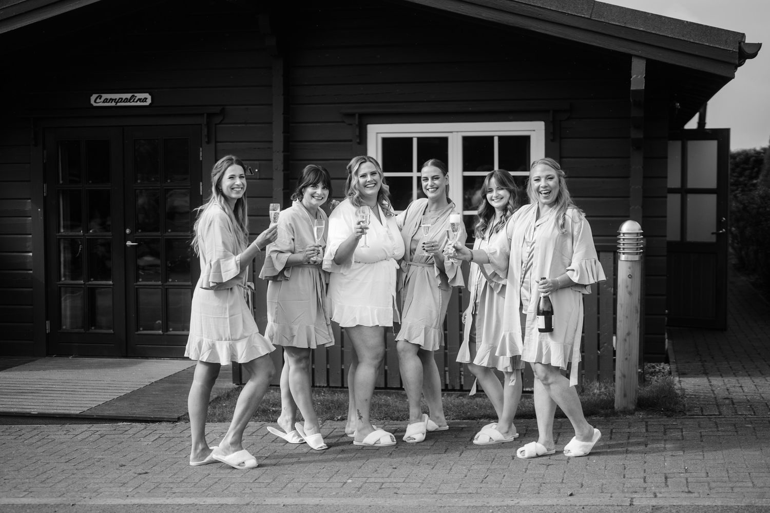 Wedding party in light colored outfits poses casually outside rustic venue.