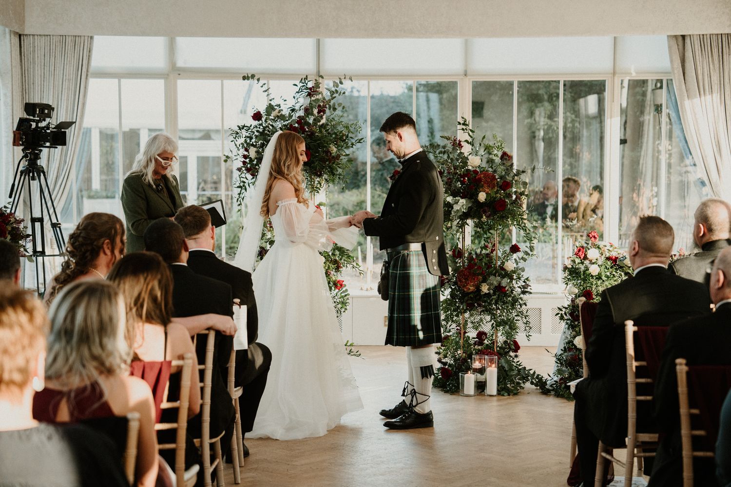 A wedding ceremony takes place in a bright conservatory decorated with red and white floral arrangements.