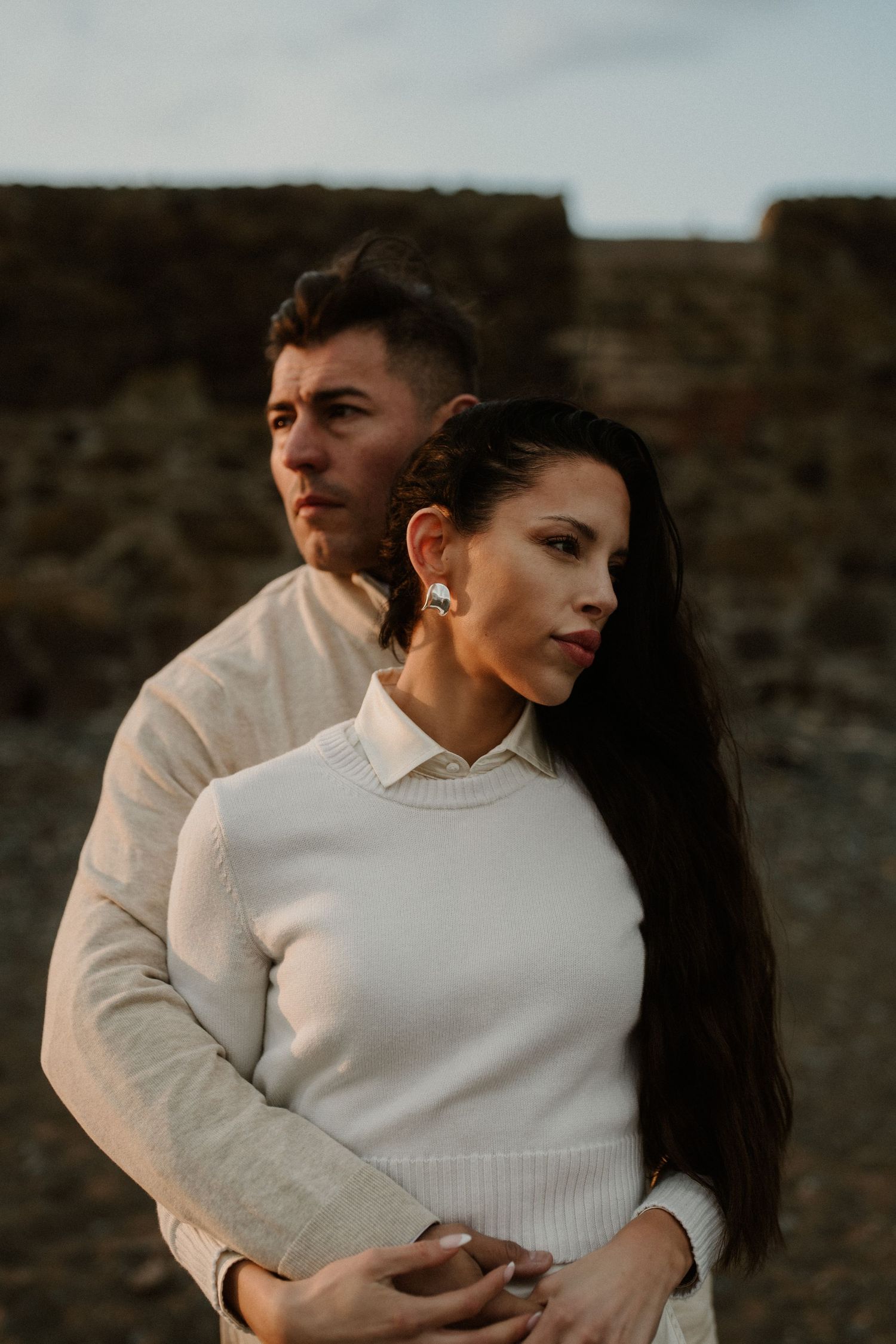 Stylish couple in white sweaters pose against coastal cliffs at sunset.