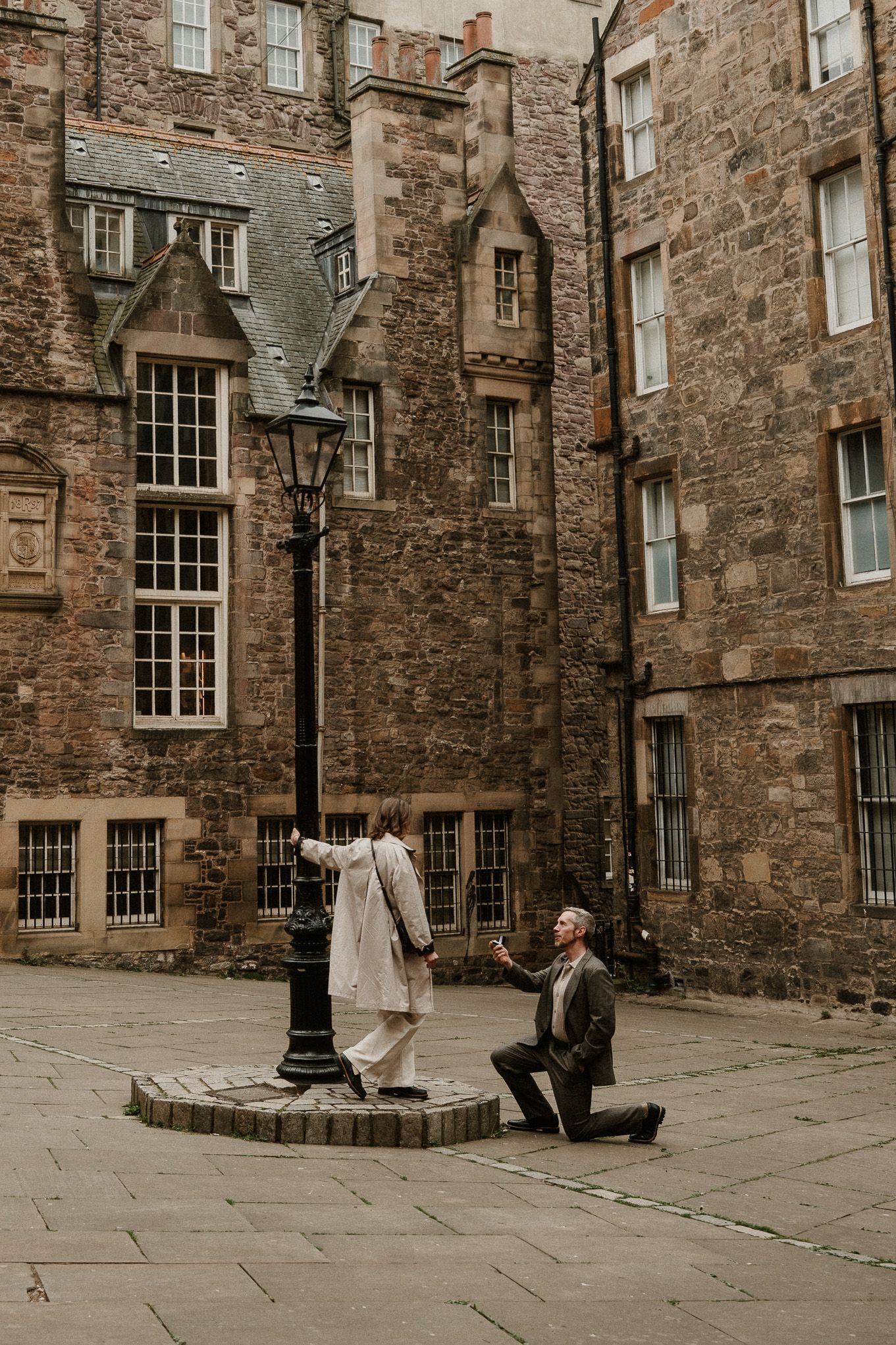 Man proposes on one knee to partner near vintage lamppost in historic Edinburgh stone courtyard.