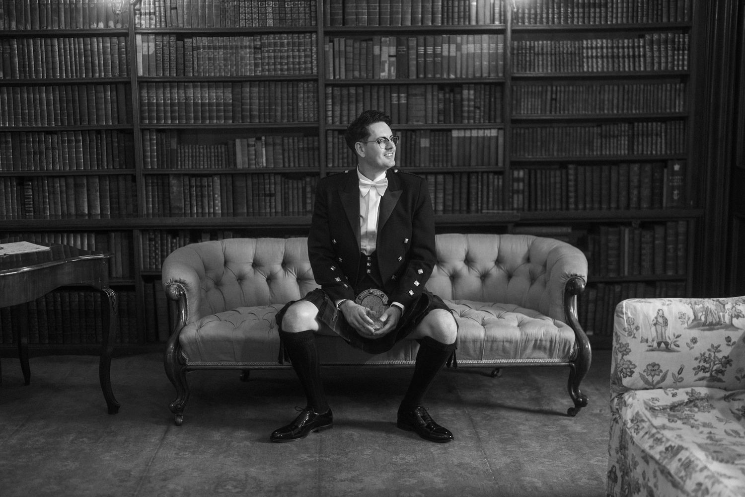 Black and white photo of a man in a kilt and jacket sitting on a tufted sofa in a classic library with floor-to-ceiling bookshelves.