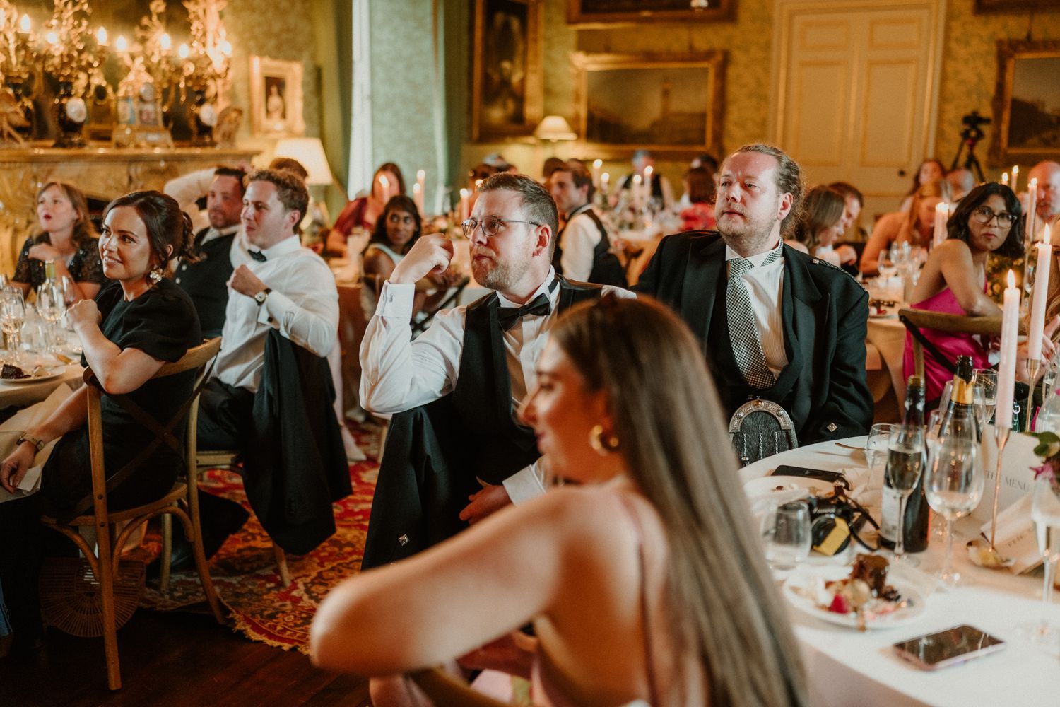 Formally dressed wedding guests seated at candlelit tables in an elegant historic banquet hall, with a woman in the foreground looking on.