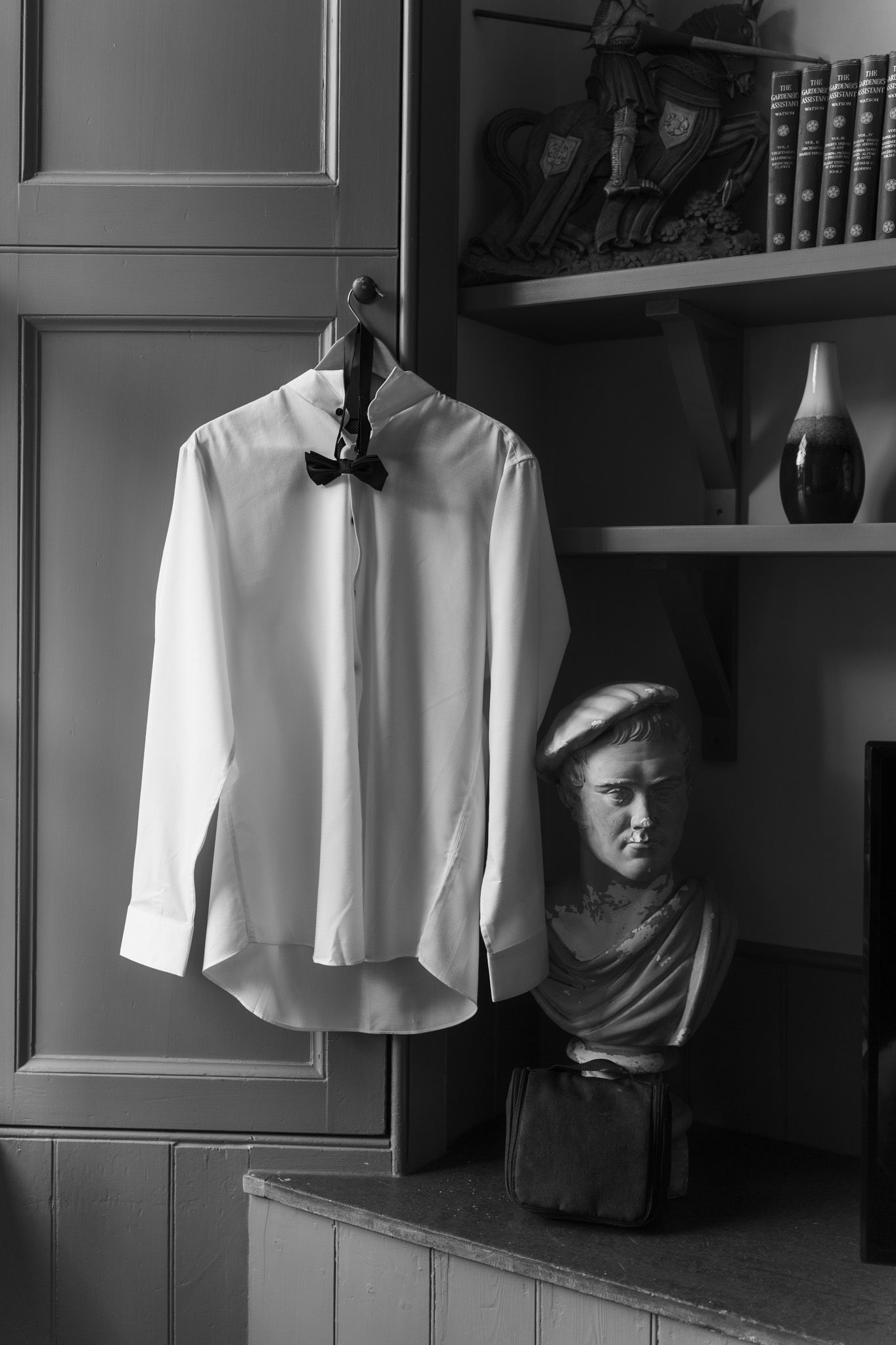 White dress shirt with a black bow tie hanging on a cabinet door beside a classical bust and bookshelf in a black and white photo.