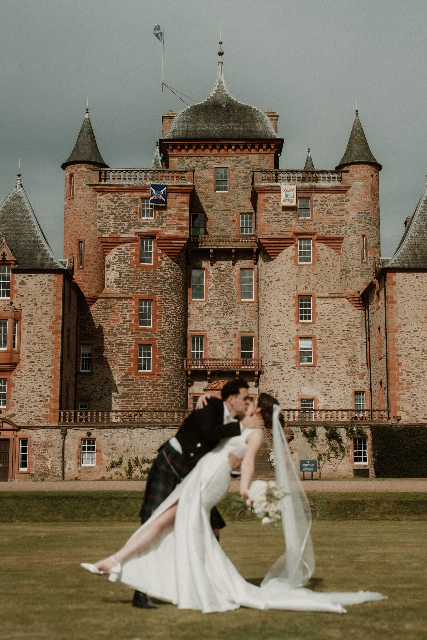 Bride and groom in a Scottish kilt share a dramatic dip kiss on the lawn in front of a red sandstone castle with turrets at a wedding.