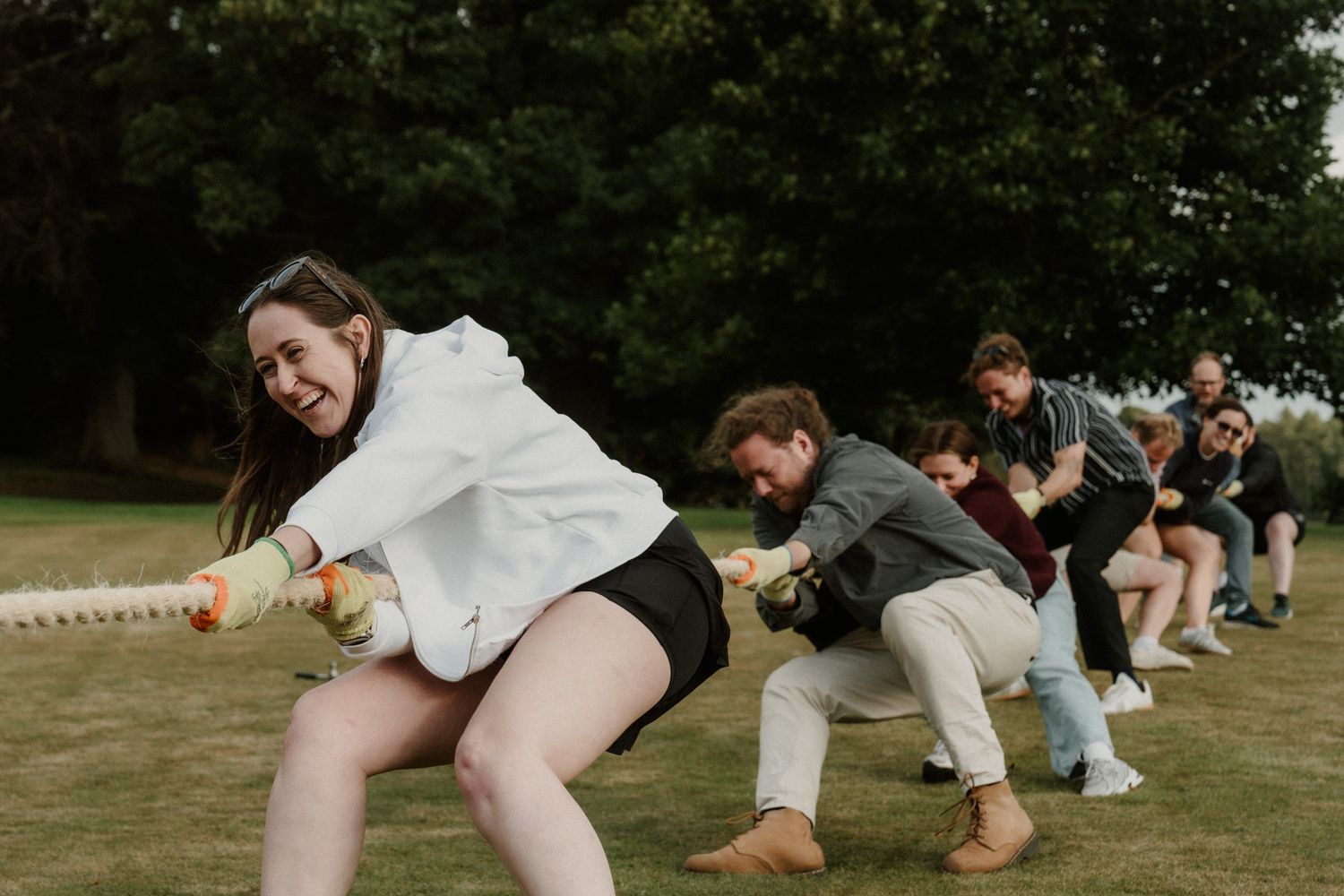 A smiling woman in a white hoodie pulls a rope with a group of people in a lively tug of war game on a grassy lawn under tall trees.