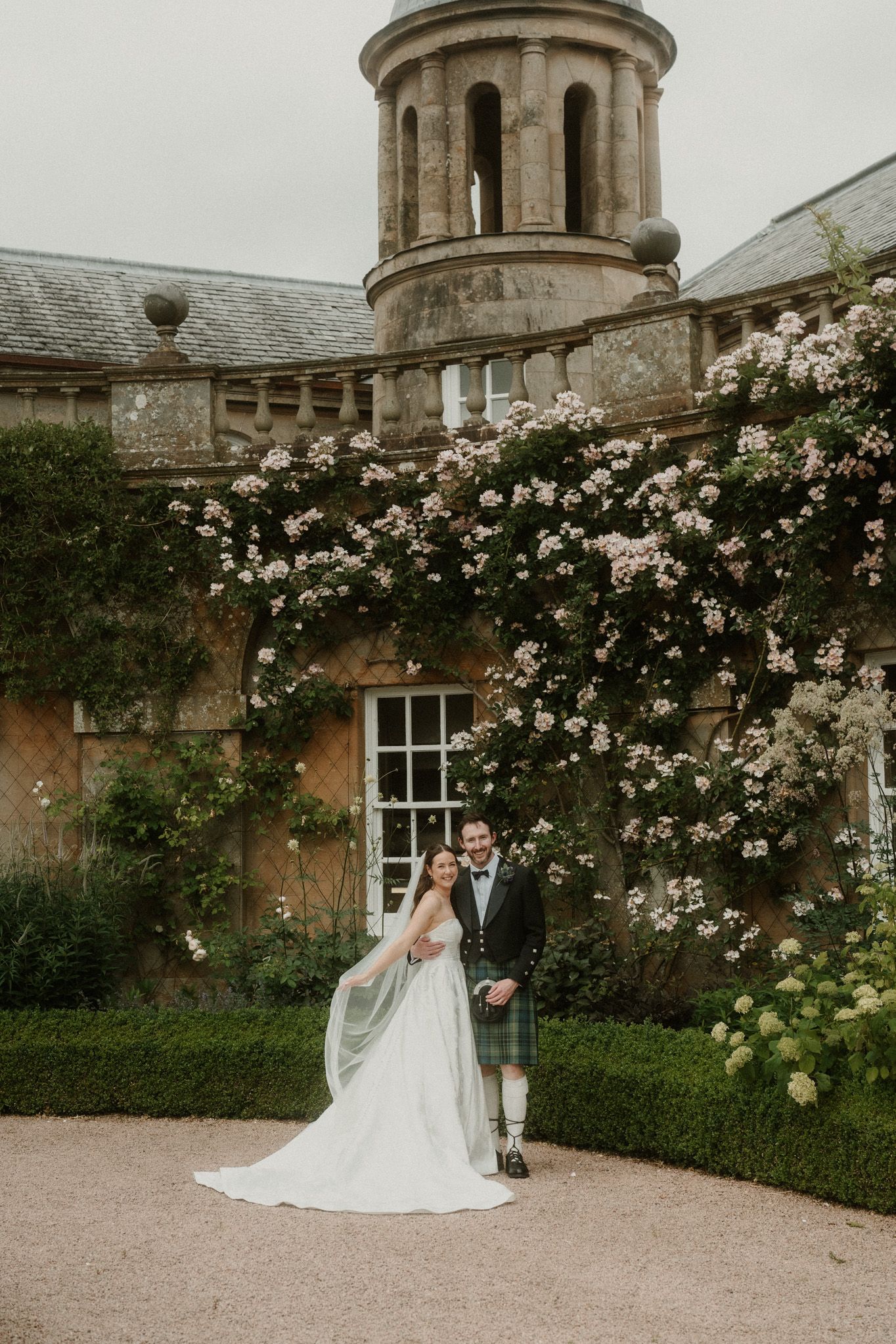 Bride and groom pose before a Scottish castle adorned with climbing pink roses, groom wearing traditional tartan kilt.