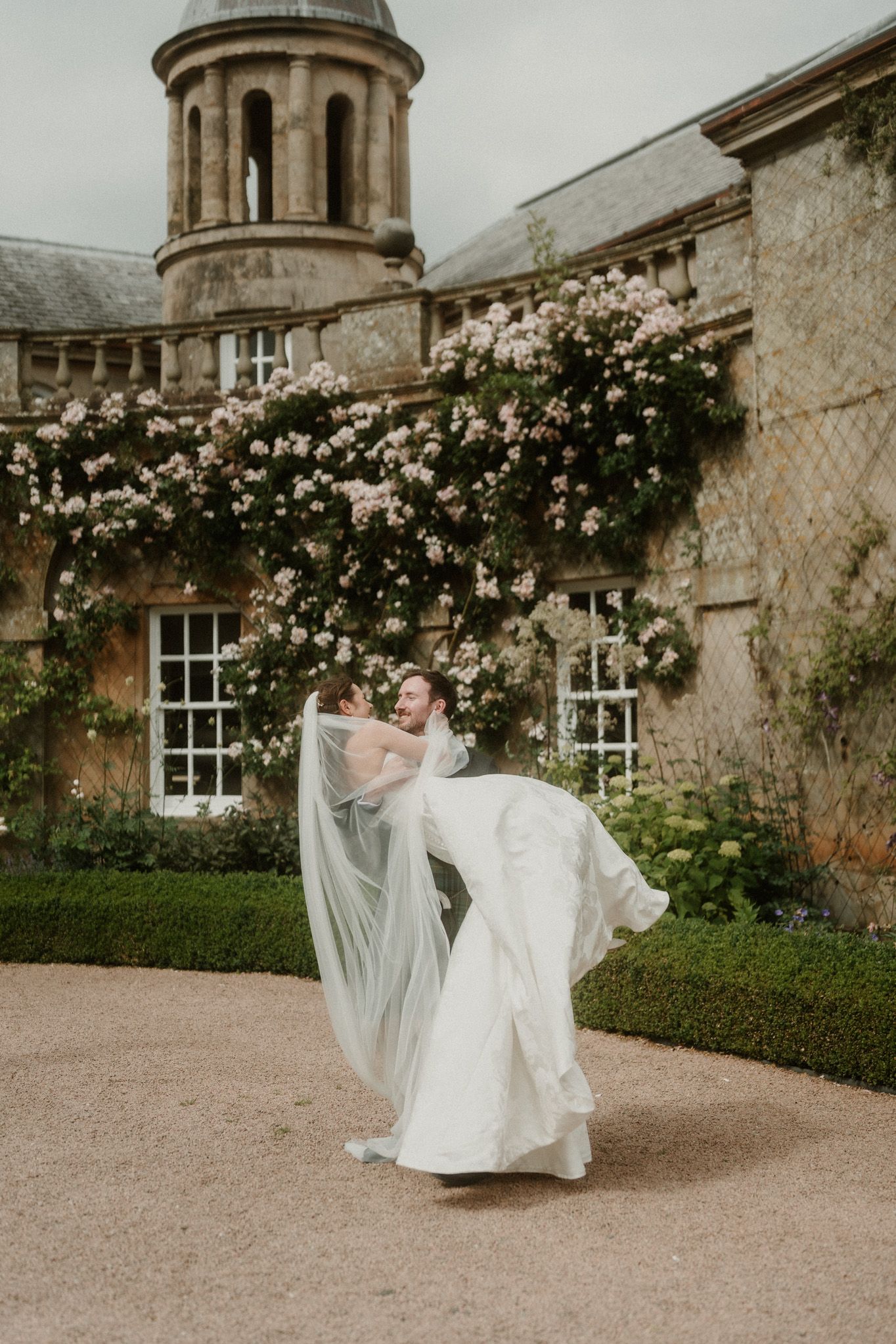 Bride in flowing white gown and veil twirls outside a historic manor adorned with climbing pink roses.