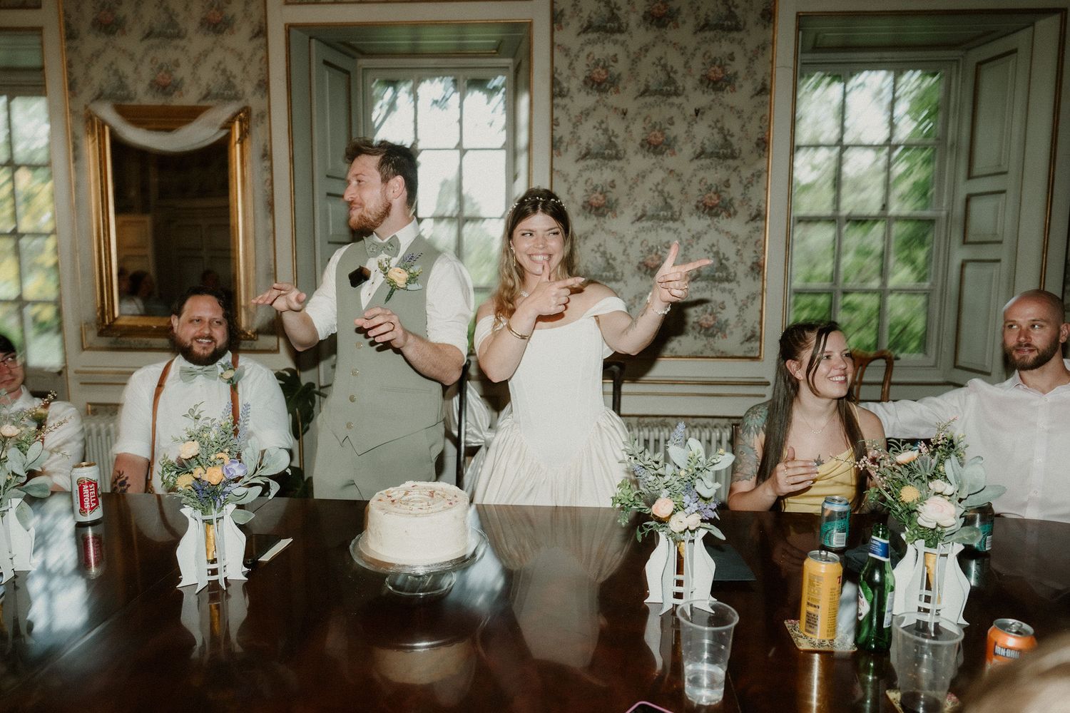 Bride and groom laughing during wedding reception speech at elegant venue with floral centerpieces and wedding cake.