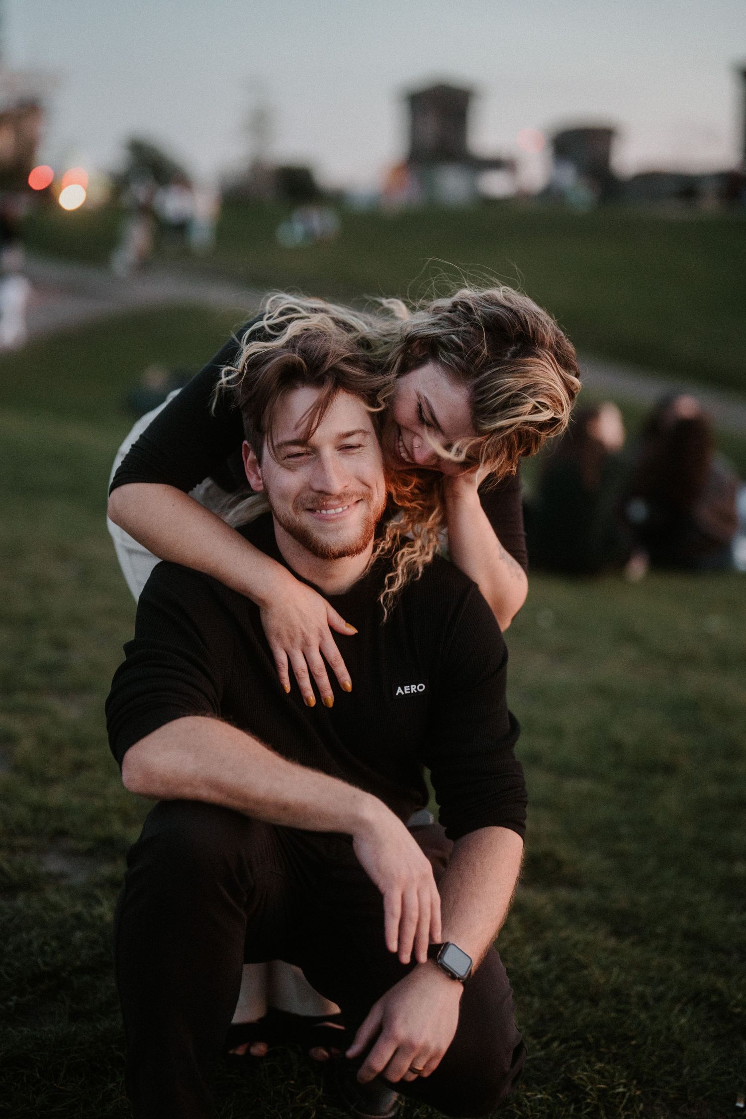 Smiling man crouching on grass while woman leans over his shoulders hugging him at dusk in a park.