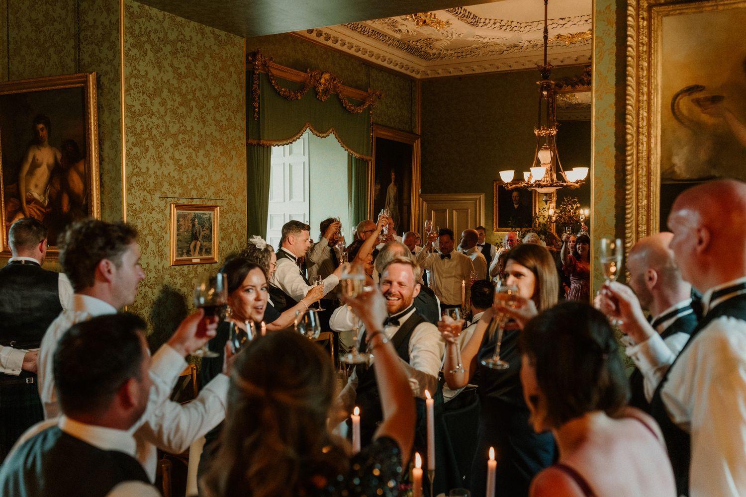 Guests in formal attire raise glasses and celebrate at a candlelit dinner in an elegant historic room with green walls and ornate details.