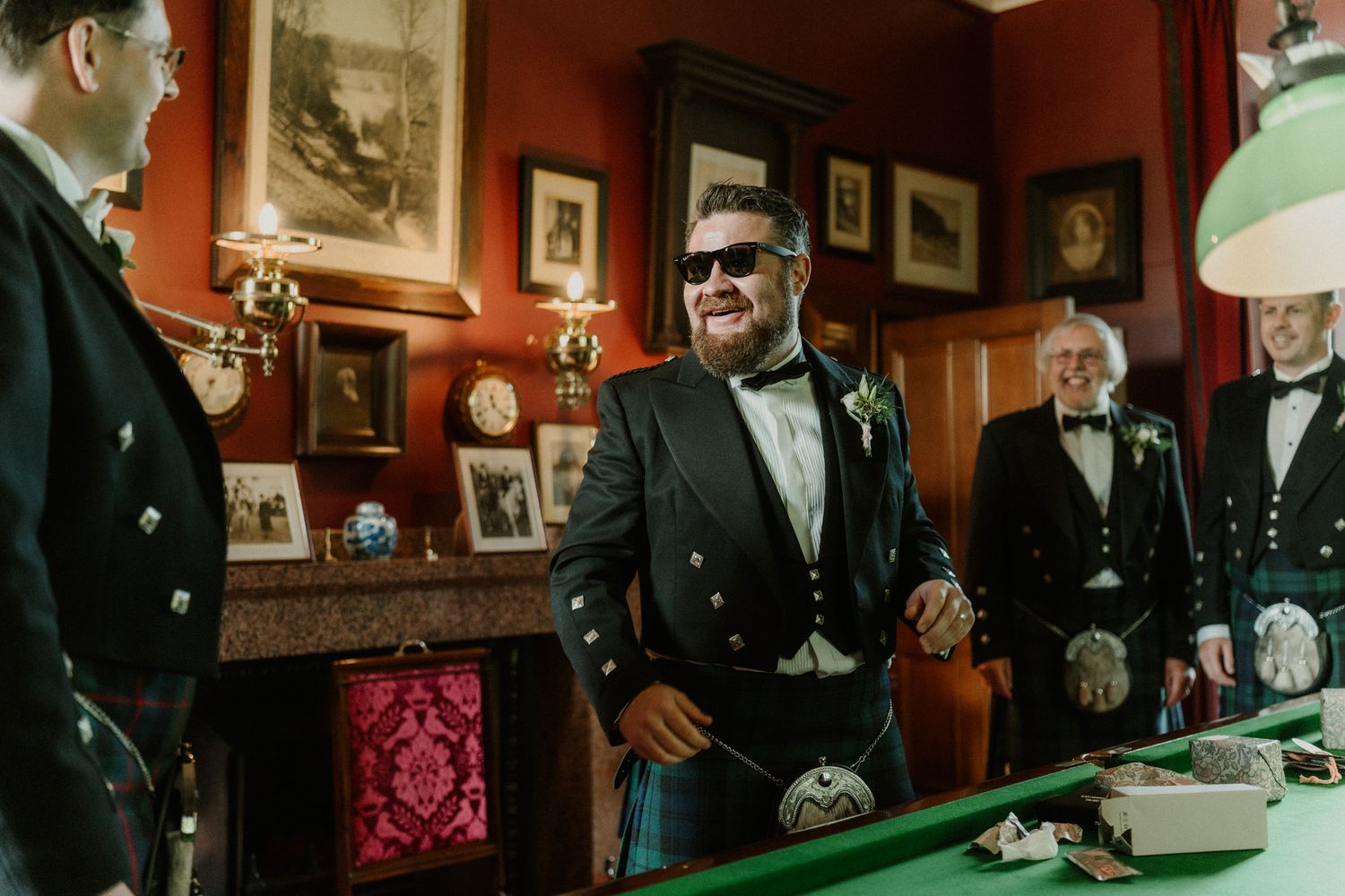 Groomsman in sunglasses and Scottish kilt formalwear smiles beside a billiards table while other kilted men chat in a wood-paneled room.