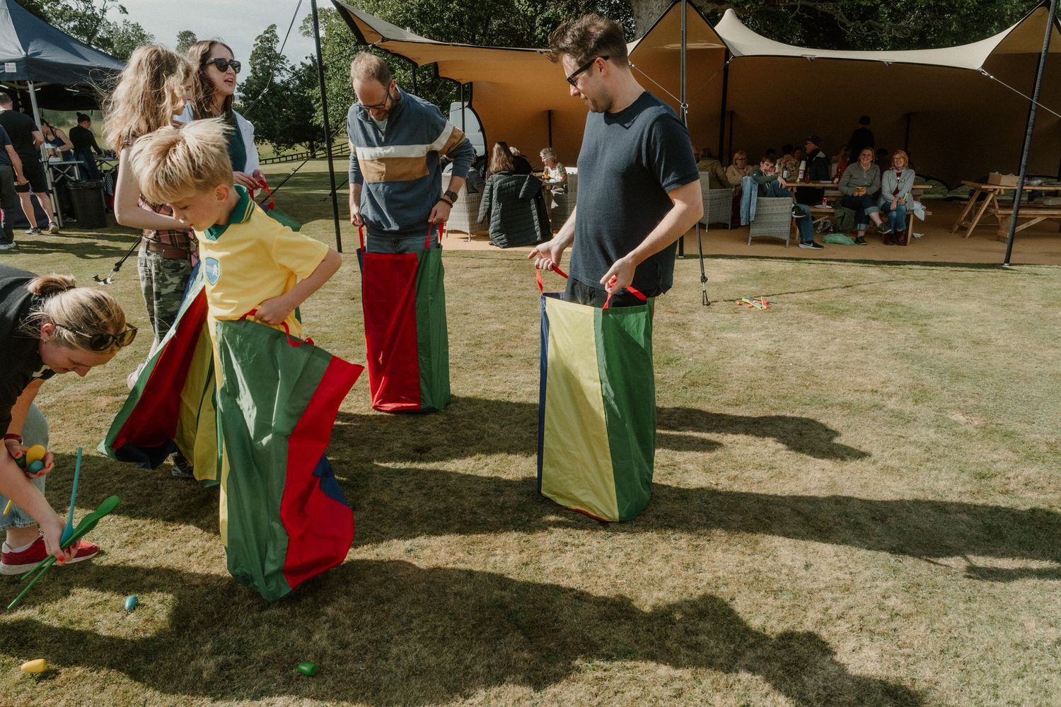 Children and adults compete in a sack race on a grassy lawn at an outdoor festival, with tents and spectators in the background.