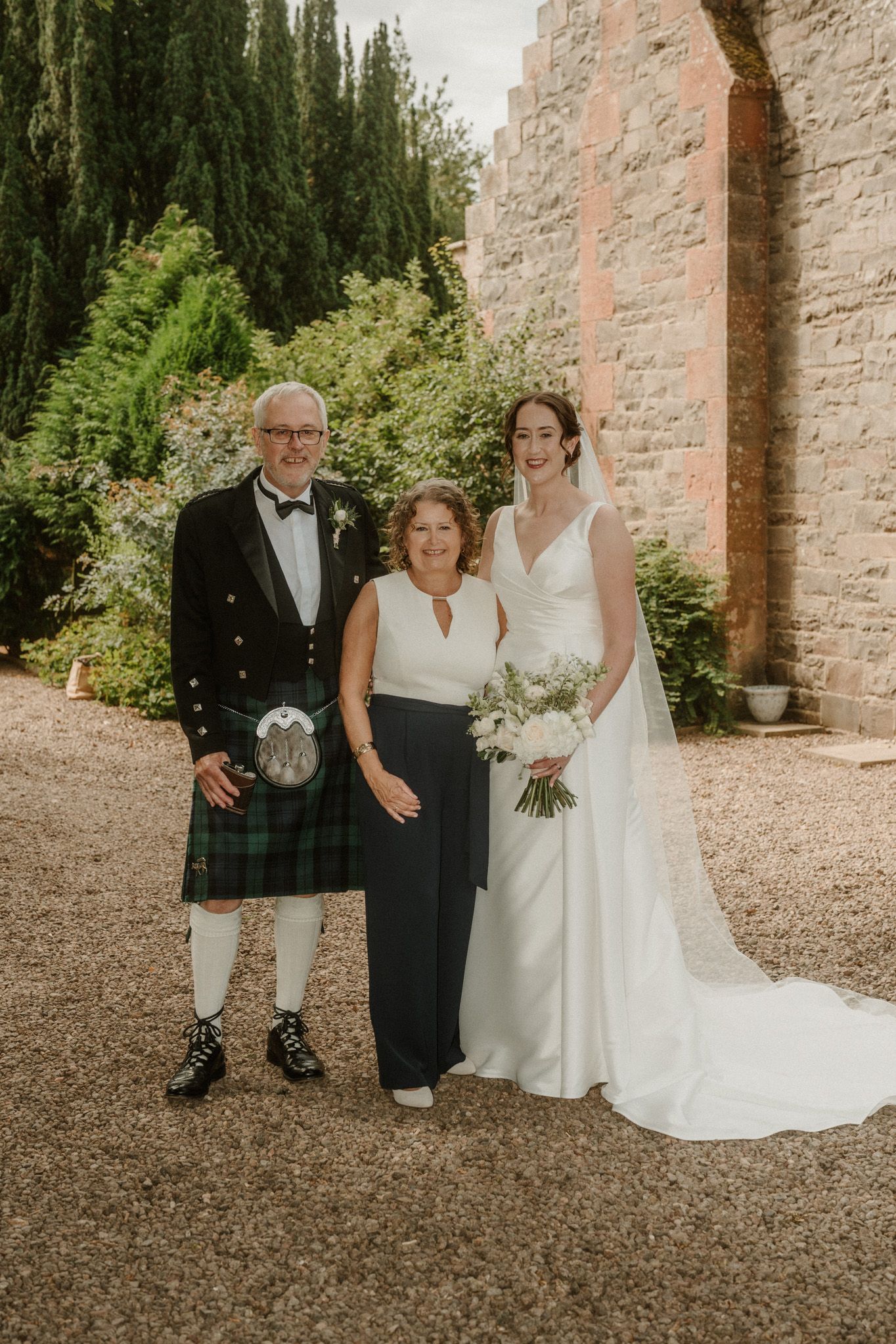 A bride in a white gown and veil holding a bouquet poses with a man in a kilt and a woman in black and white outside a stone building.
