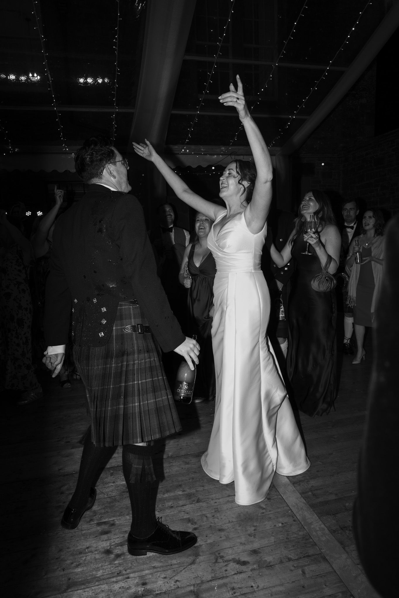 Bride in a satin gown dances with arms raised beside a man in a kilt on a dance floor at a lively evening wedding reception.