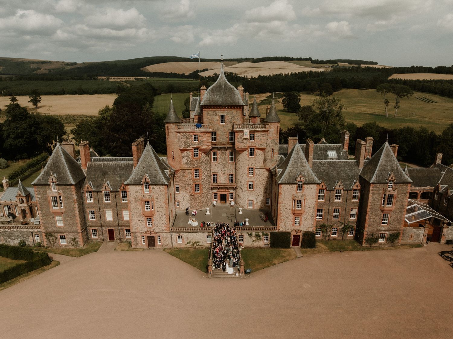 Aerial view of a historic stone castle with turrets surrounded by green countryside, with a wedding group gathered on the front lawn.