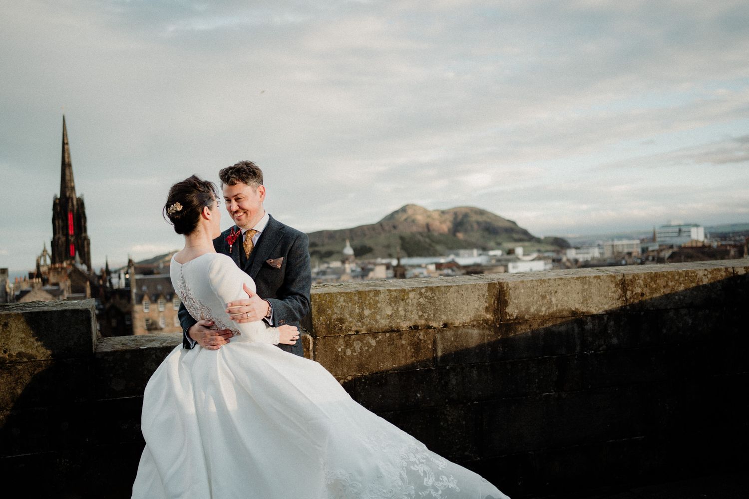 Couple embraces on castle walls with Edinburgh's iconic skyline in background.