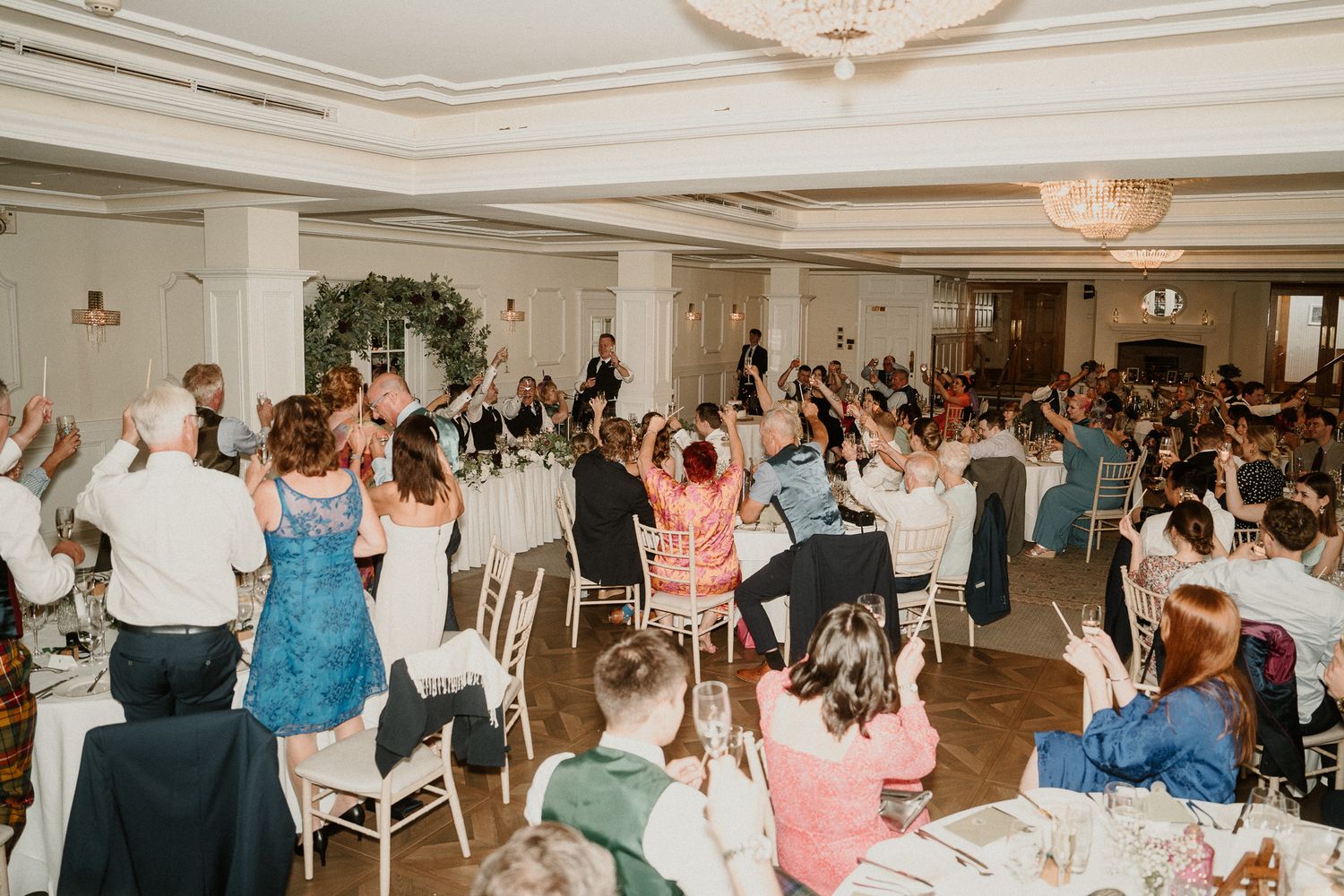 Wedding reception guests raising their hands in celebration during a joyous moment at an elegant indoor venue.