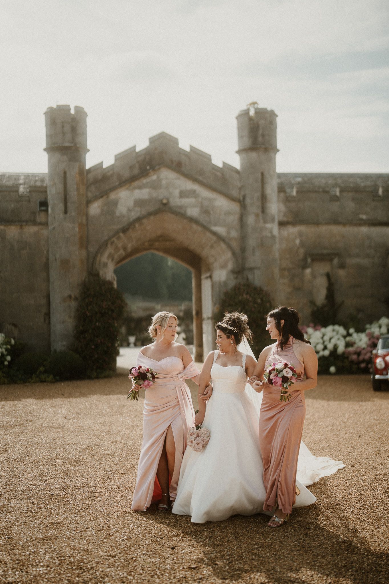 Bridesmaids in blush dresses help arrange bride's gown outside castle entrance.